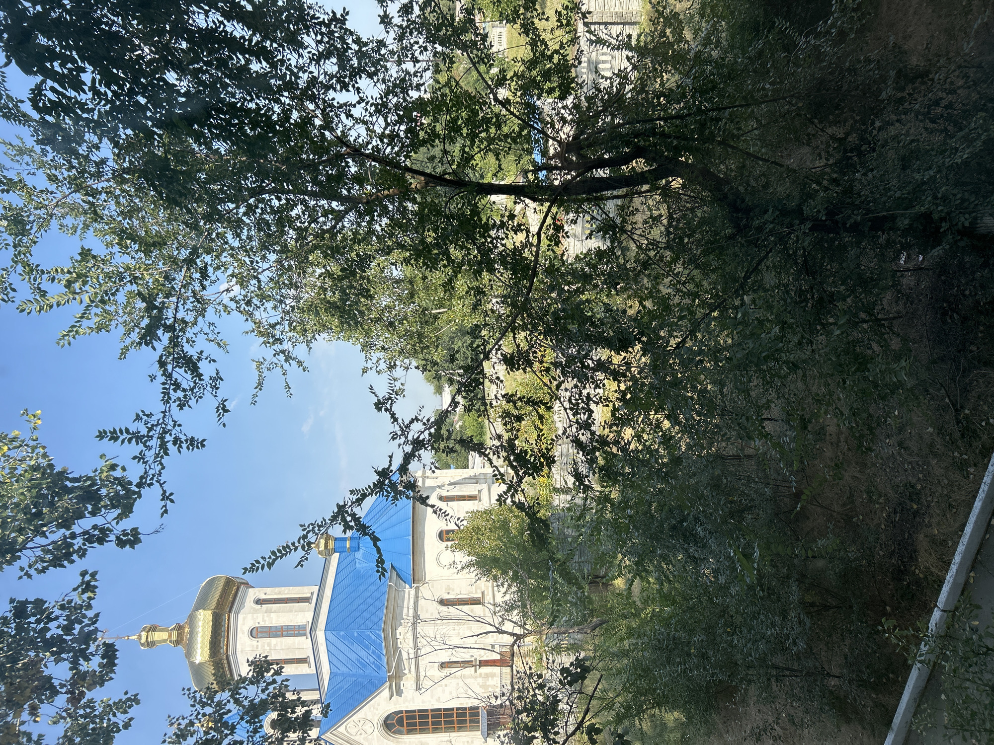 The image shows an exterior view of a white church with a bright blue roof and a golden dome, situated in the city of Basarabeasca, Moldova. The church is partially obscured by lush green foliage from trees in the foreground. The sky is clear and blue. The church has tall, arched windows with orange-tinted glass. A stone wall with arched details is visible behind the trees, and further in the background, other buildings and trees are present. The scene appears to be a sunny day.