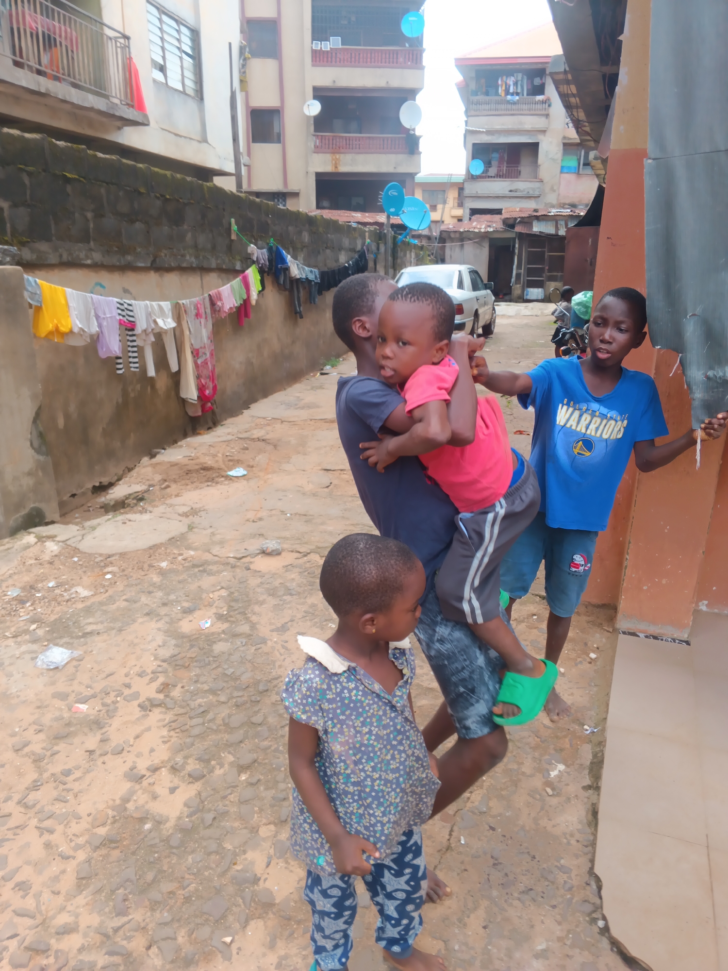 Four Black children are gathered on a dusty, uneven ground in front of a building. The building appears to be a multi-story residential structure with balconies. Laundry is hanging on a line in front of a lower wall. The sky is overcast, suggesting a cloudy day.
The most prominent subject is a group of three children in the center. A slightly older boy is holding a younger child in his arms, while another child stands to his right. The child being held is wearing a pink t-shirt and grey shorts with white stripes. He is looking towards the viewer with a serious expression. The boy holding him is wearing a dark t-shirt and jeans, and he is also looking at the camera. The third child, on the right, is wearing a blue t-shirt with the text "GOLDEN WARRIORS" and a basketball logo. He is looking off to the side. To the left of this group, a young girl in a patterned dress and blue-and-white patterned leggings is looking down.
The scene appears to be a residential area in Onitsha, Nigeria. The ground is a mix of dirt and small stones, with some litter visible. The buildings suggest an urban environment. The overall impression is one of everyday life in a community.