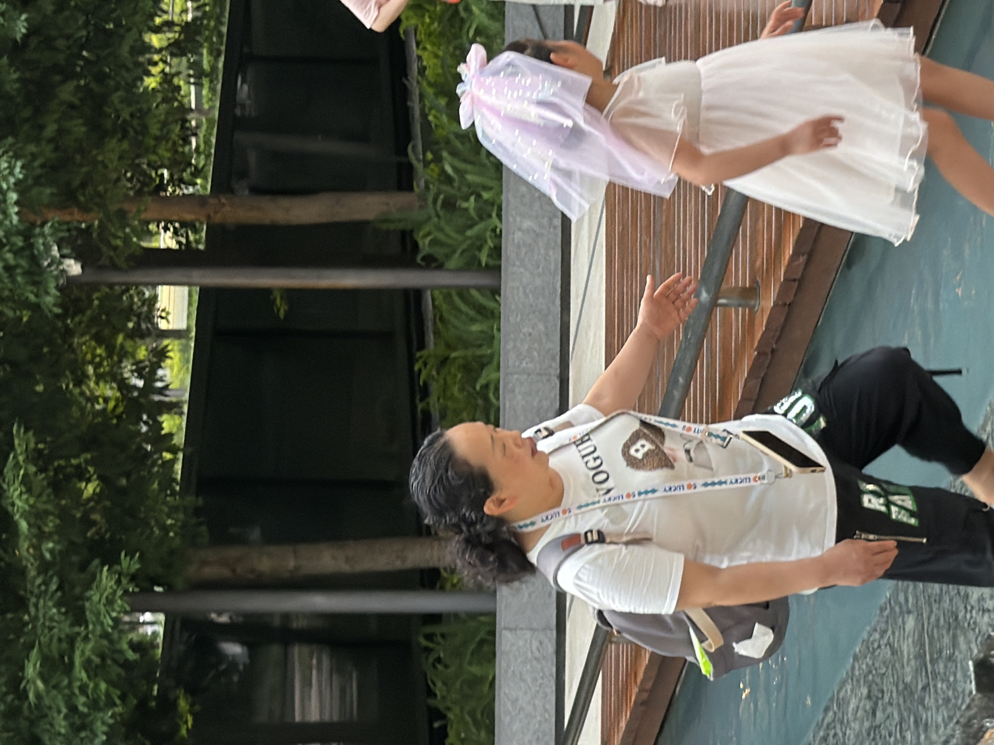 This outdoor, eye-level, full-length shot depicts an adult woman and a young girl walking in what is identified as Yuzhong District, China. The woman, positioned left of center, has dark hair pulled back and is wearing a white short-sleeved t-shirt with "VOGUE" and a brown graphic, black pants with partially visible green lettering ("RU FAIR") and a zipper detail on the left thigh. A grey backpack with light straps is on her back, and a gold-colored smartphone hangs from a "LUCKY" branded lanyard around her neck. Her left arm is extended forward, palm open, seemingly gesturing towards the child. Her mouth is slightly ajar, and her gaze is directed towards the child.
The young girl, positioned right of center and slightly ahead of the woman, is wearing a white, sheer, short-sleeved dress. On her head, she has a translucent light pink/purple veil-like hair accessory with a bow at the top and shimmering elements within the fabric. Her back is mostly to the camera as she walks forward.
The foreground features a dark blue-green water body or shallow pool that partially obscures the woman's lower legs. A wooden walkway or bridge with horizontal planks and a dark metal railing runs diagonally from mid-left to mid-right. Beyond this structure, a grey concrete wall and lush green foliage are visible. The background consists of tall trees with dense green canopies and dark trunks, interspersed with dark, possibly metal, barrier walls. A vertical dark grey pole stands in the mid-background. The lighting is bright and even, suggesting daytime.