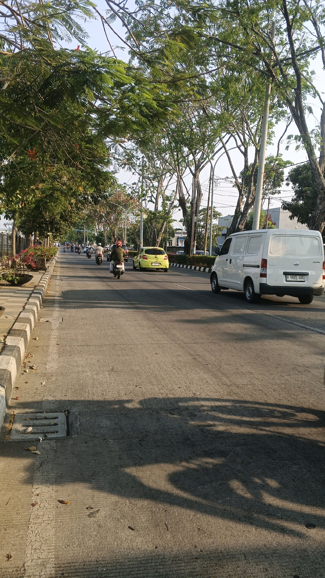 This image depicts a street scene in Semarang, Indonesia. The perspective is from street level, looking down a paved road lined with trees on the left and a sidewalk. Several motorcycles and a yellow car are traveling down the road. A white van is also present on the right side of the road. The sun is casting long shadows across the road, indicating it is likely late afternoon or early morning. The area appears to be a moderately busy urban environment with vegetation and street infrastructure.