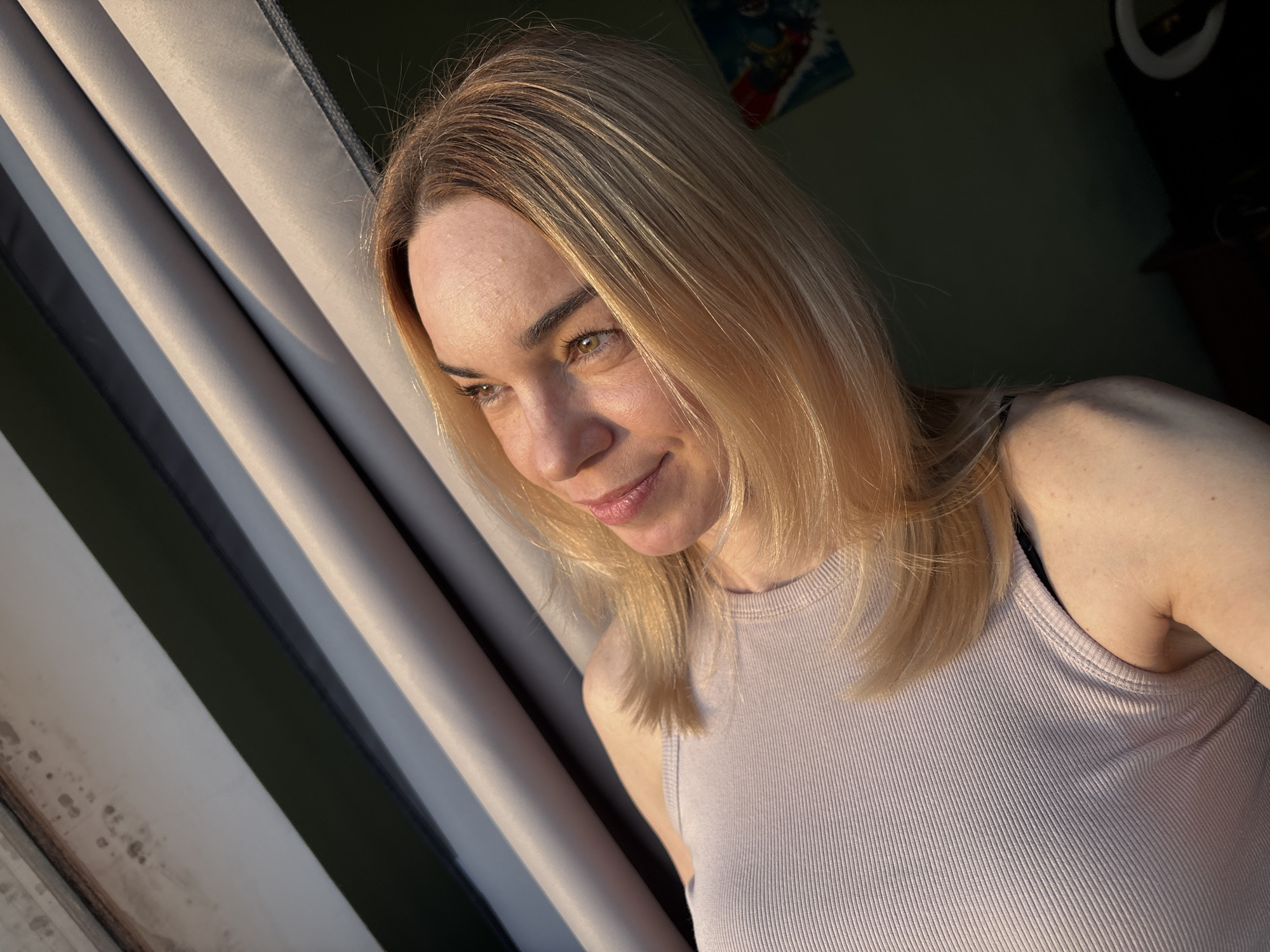 A medium close-up portrait features a woman with fair skin and shoulder-length blonde hair, captured indoors. She is positioned slightly to the right of the frame, looking towards the left with a gentle, soft smile and light-colored eyes, possibly green or hazel. Golden sunlight illuminates the right side of her face and hair, creating warm highlights and subtle shadows. She is wearing a light purple or pink ribbed tank top.
The background on the left consists of vertical blinds or curtains in varying shades of grey and beige, creating a striped pattern with alternating bands of light and shadow, suggesting direct sunlight streaming in from the left. To the right, behind her head and shoulder, the background is darker, indicating an interior room. A hint of a light source, possibly a ring light, and a colorful, unfocused image (possibly a poster) are visible in the darker background. The overall mood is calm and contemplative, likely taken during late afternoon or early morning due to the warm, directional light. There is no visible text in the image. The location is unknown, in Russia.