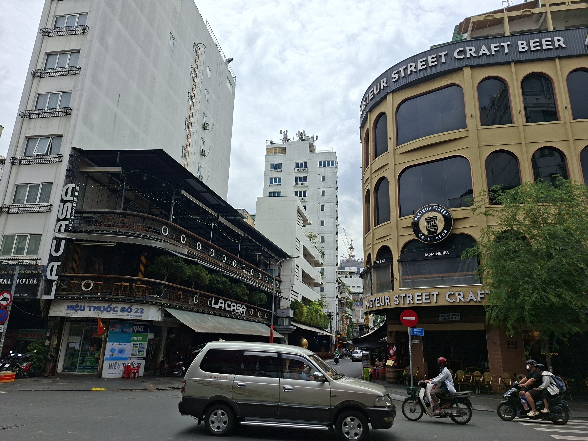 The image depicts a street scene in Thủ Đức City, Vietnam, on a cloudy day. In the foreground, a silver minivan with the license plate "51H-07.007" is driving on the right side of the road. To the right of the minivan, two motorcycles are also traveling. The first motorcycle is ridden by a person wearing a red helmet and a white shirt, and the second motorcycle carries two individuals, one wearing a black helmet and the other a backpack.
The background features several buildings. On the left, a tall, white building with multiple floors and balconies is visible. Adjacent to it is a dark, multi-story building with a balcony overlooking the street, adorned with string lights. A sign on this building reads "LA CASA". Further down the street, another tall, white building stands, topped with antennas.
On the right side of the image, a prominent, tan-colored building with arched windows dominates the scene. A large sign on this building reads "PASTEUR STREET CRAFT BEER". Below this sign, another sign indicates "JASMINE IPA". Green trees partially obscure the lower portion of this building.
Signs for businesses like "HIỆU THUỐC SỐ 22" (Pharmacy Number 22) and "MASSAGE" are also visible on the street level. The overall environment suggests a bustling urban area with a mix of commercial establishments and residential buildings.