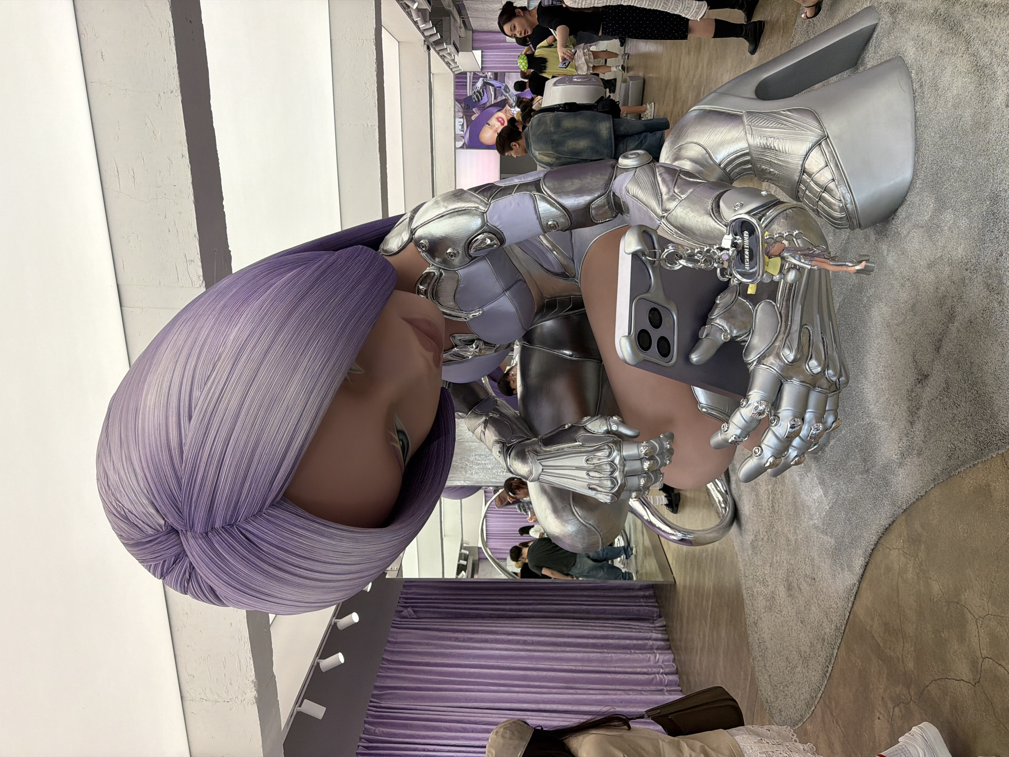 The image depicts a large, futuristic sculpture of a female figure with silver, robotic armor and a purple, turban-like head covering. The figure holds a mobile phone with a keychain attached to its case. The sculpture is located inside a retail space, likely a fashion store, in Seoul, South Korea. In the background, there are people browsing through clothing racks and interacting with the merchandise, as well as some promotional posters. The retail space has purple curtains, white walls, and a gray carpet. The overall setting indicates a modern, design-conscious retail environment.