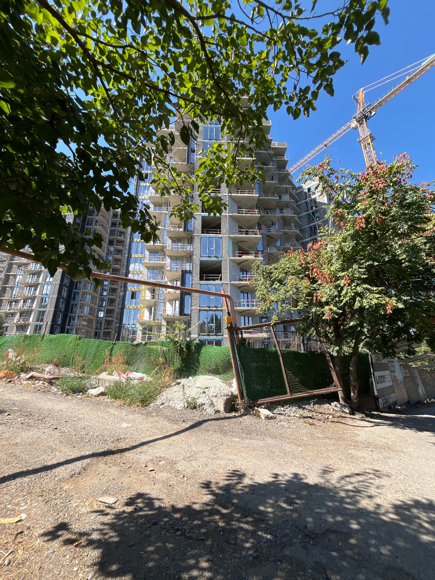This is a ground-level, eye-level shot of a high-rise apartment building under construction in Tbilisi, Georgia. The building is a multi-story concrete structure with visible scaffolding and many windows, some of which are covered. A yellow construction crane is visible in the background, soaring above the building against a clear blue sky. In the foreground, there is a dirt path, a green fence, and some overgrown vegetation. A rusty metal gate stands partially open, leaning against the fence. The scene is illuminated by bright sunlight, casting distinct shadows from trees onto the ground. The overall impression is one of ongoing urban development.