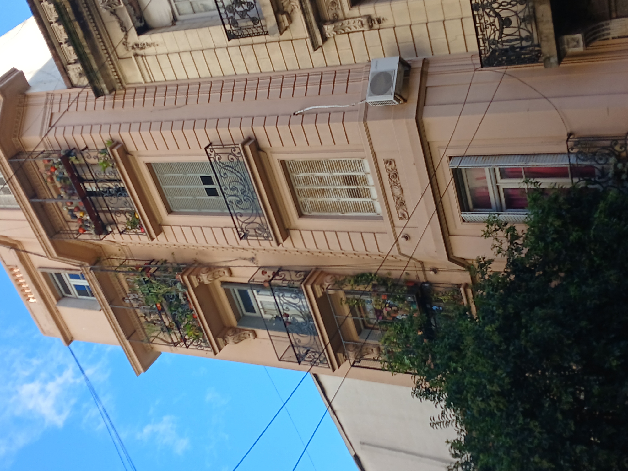 The image displays a multi-story building facade, captured from a low angle and significantly rotated clockwise. This perspective offers a view up towards the building against a bright, clear daytime sky with scattered clouds, characteristic of an urban setting, likely in Buenos Aires, Argentina.
The building is painted in a light peachy-pink or salmon color and features a classic architectural style with several visible floors. Each floor is adorned with distinct windows and decorative black wrought-iron balconies or railings. Many windows are fitted with traditional louvered wooden shutters, some of which are closed, while others are partially or fully open.
Numerous balconies and window sills are decorated with green plants and colorful flowers in pots, suggesting a well-maintained and vibrant environment. On an upper floor towards the right, a white, rectangular air conditioning condenser unit is mounted on the exterior wall. The facade also features subtle architectural details such as horizontal banding, vertical fluting, and decorative moldings around windows and below balconies.
In the lower right portion of the frame, a large, dark green tree or bush partially obscures the building's base. The bright blue sky in the background shows a few wispy white clouds. Several thin, dark electrical or utility wires stretch diagonally across the sky in the lower-left section of the image. The scene is illuminated by strong daylight, indicating a clear, sunny day. No people or visible text are present in the image.
