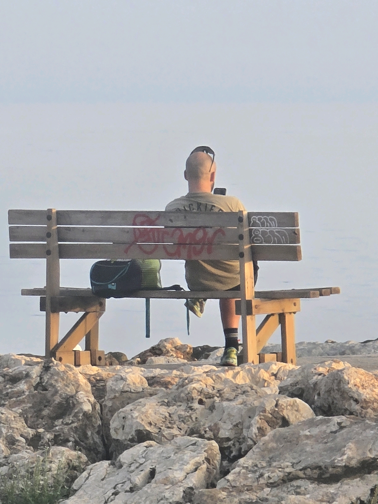 A bald man with sunglasses on his head sits on a wooden bench facing a body of water. He is wearing a green t-shirt, dark shorts, striped socks, and athletic shoes. The man is looking down at a mobile phone he is holding in his right hand. A dark green and blue backpack rests on the bench to his left. The bench is made of weathered wood and has graffiti on it, including red spray paint on the backrest and white outlined figures on the right side. The bench is situated on a rocky surface overlooking a calm body of water, possibly the Mediterranean Sea. In the foreground, large, light-colored rocks are visible, with some sparse green vegetation. The sky is clear and a pale blue. This scene is consistent with a location along the coast of Málaga, Spain, likely an area with public seating overlooking the sea.