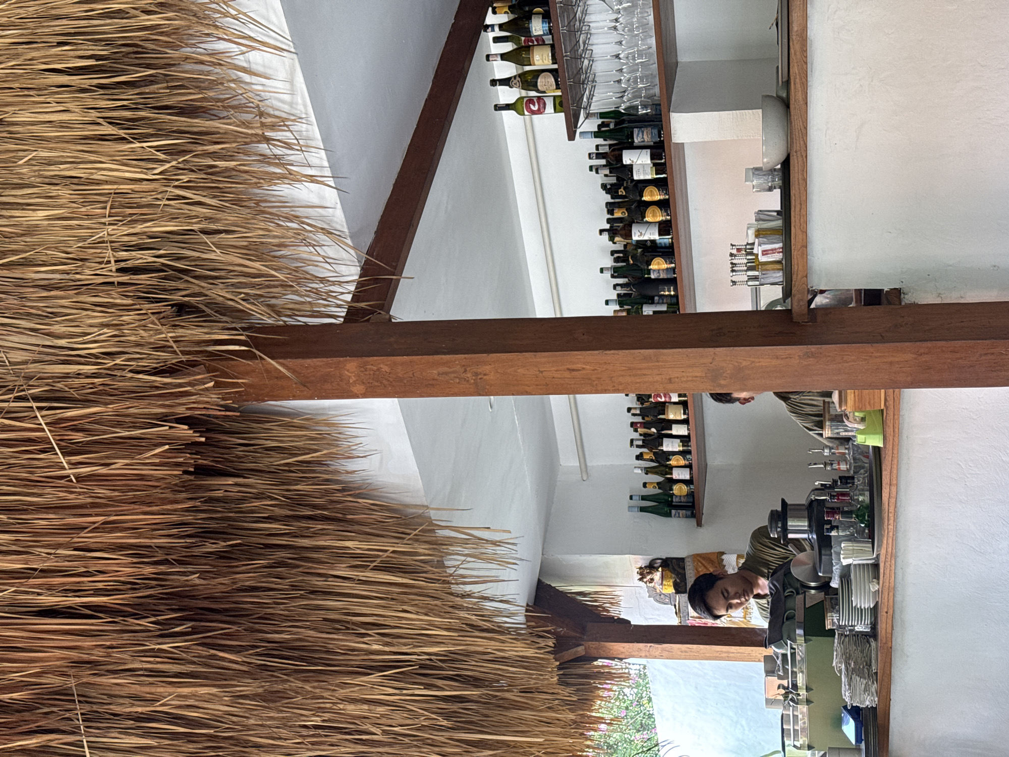 The image shows the interior of a building in North Kuta, Indonesia. The left side of the frame is dominated by a thick layer of thatching, likely part of the roof. A dark wooden beam runs horizontally across the center of the image. In the background, along a white wall, there are shelves lined with wine bottles and glassware. A person is visible behind a counter. The counter area includes various bar tools and a variety of bottles and items. Part of a painted backdrop is visible in the background, showing a blue sky.