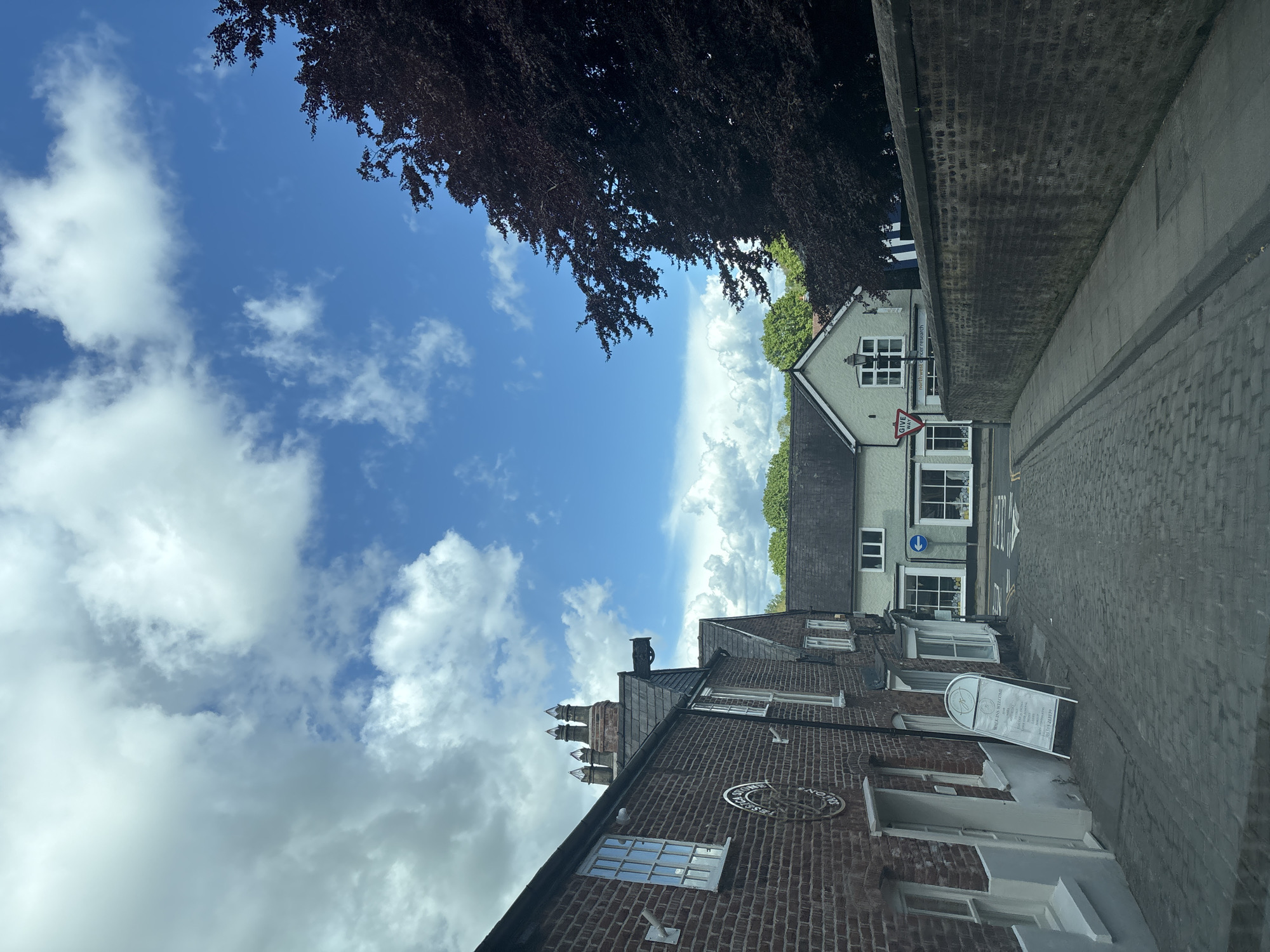 The image shows a street in Knutsford, United Kingdom. The sky is blue with scattered white clouds. A tree with dark red leaves overhangs the scene. On the right side of the street is a tall brick wall, below which is a cobblestone road. Buildings, including a light green building with a "Give Way" sign, are visible along the street. A dark brick building with decorative brickwork and a sign that reads "The Knutsford Tapestry" and an A-frame sign that reads "Watkins Welcome" are also seen.