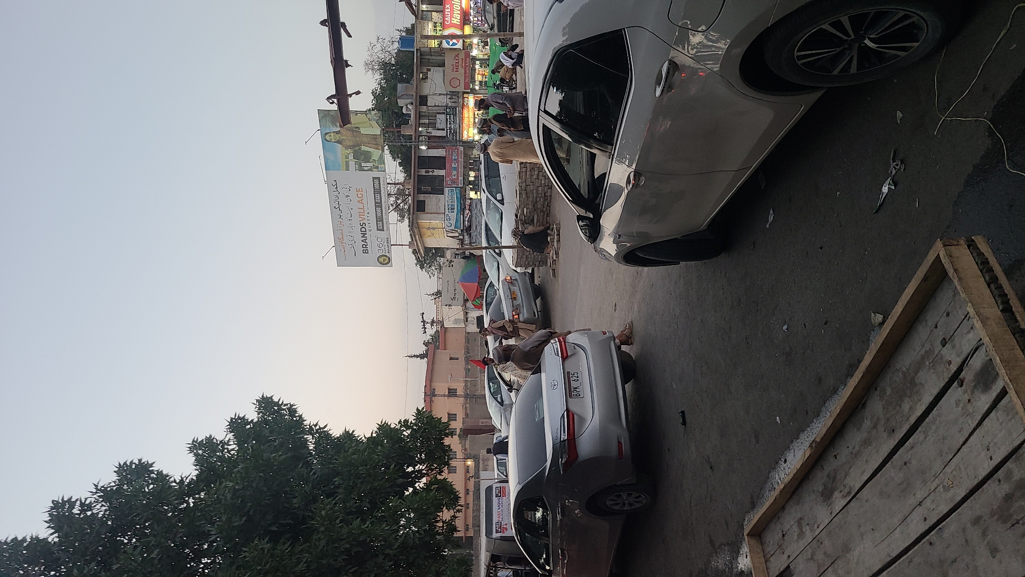 The image depicts a street in Quetta City Tehsil, Pakistan, with several parked cars along the side of the road. A few people are walking or standing near the vehicles, and some appear to be resting on one of the cars. Buildings and billboards line the street, indicating a commercial area. The sky is overcast, suggesting late afternoon or early evening. The light is fairly dim.