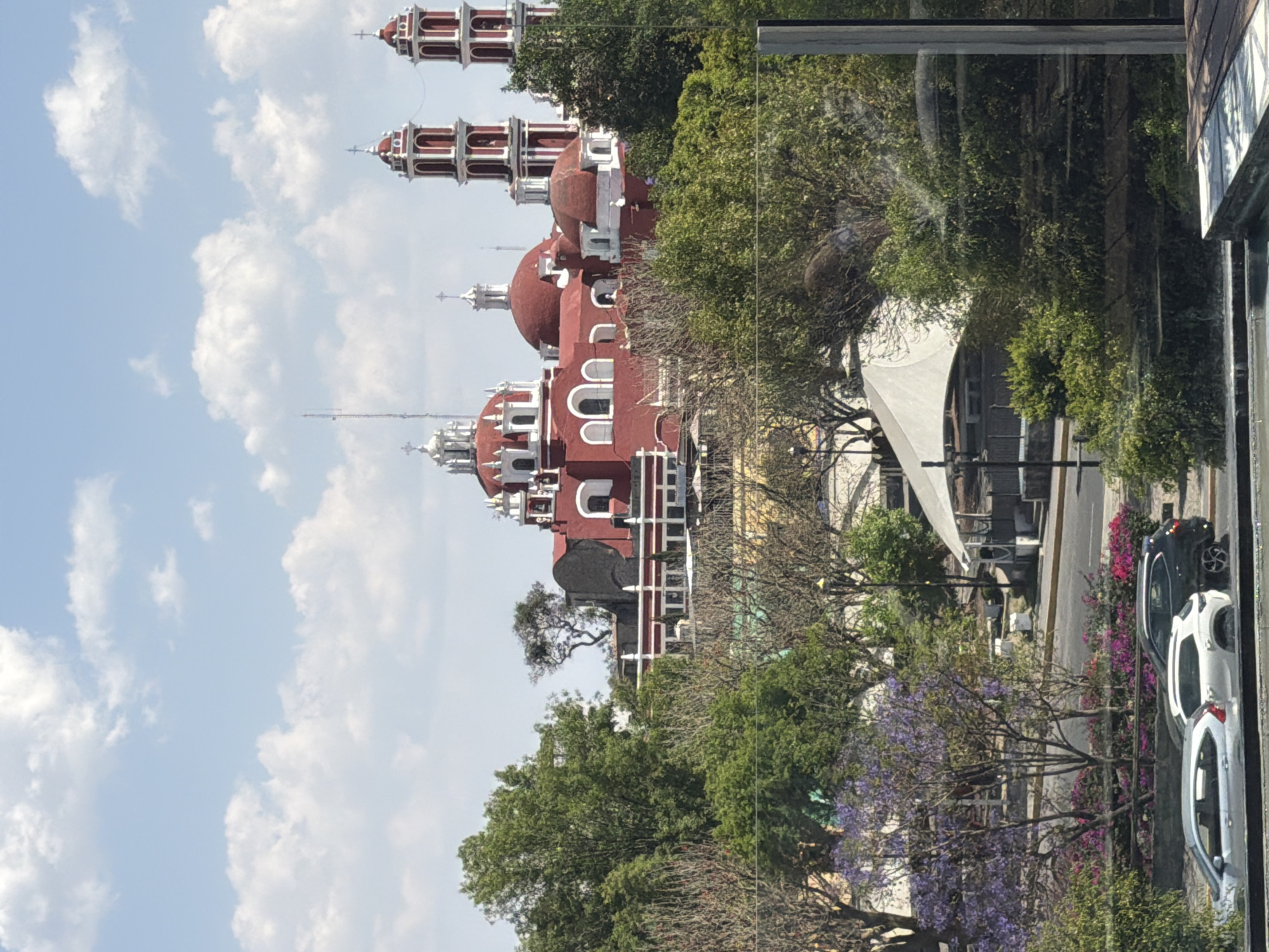 The image displays an elevated cityscape view within the city of Unknown, Unknown, under a sky with scattered white clouds. The central subject is a large religious complex, predominantly red with white architectural detailing, featuring multiple domes topped with white lanterns and crosses. Two prominent, multi-tiered bell towers, also red and white, stand on the right side of the main structure. This complex is situated amidst dense green trees and foliage, some with purple blossoms, and appears to be on a slight elevation. In the lower foreground, a paved street contains three parked vehicles: a silver sedan, a white SUV, and a dark SUV. A green median with purple-flowering trees and pink shrubs separates the street from the immediate vantage point. A large white tent-like structure is visible among the trees to the left of the main building. The bottom edge of the image shows a partial view of a reflective surface and a dark wooden or tiled platform. No individuals are visible.