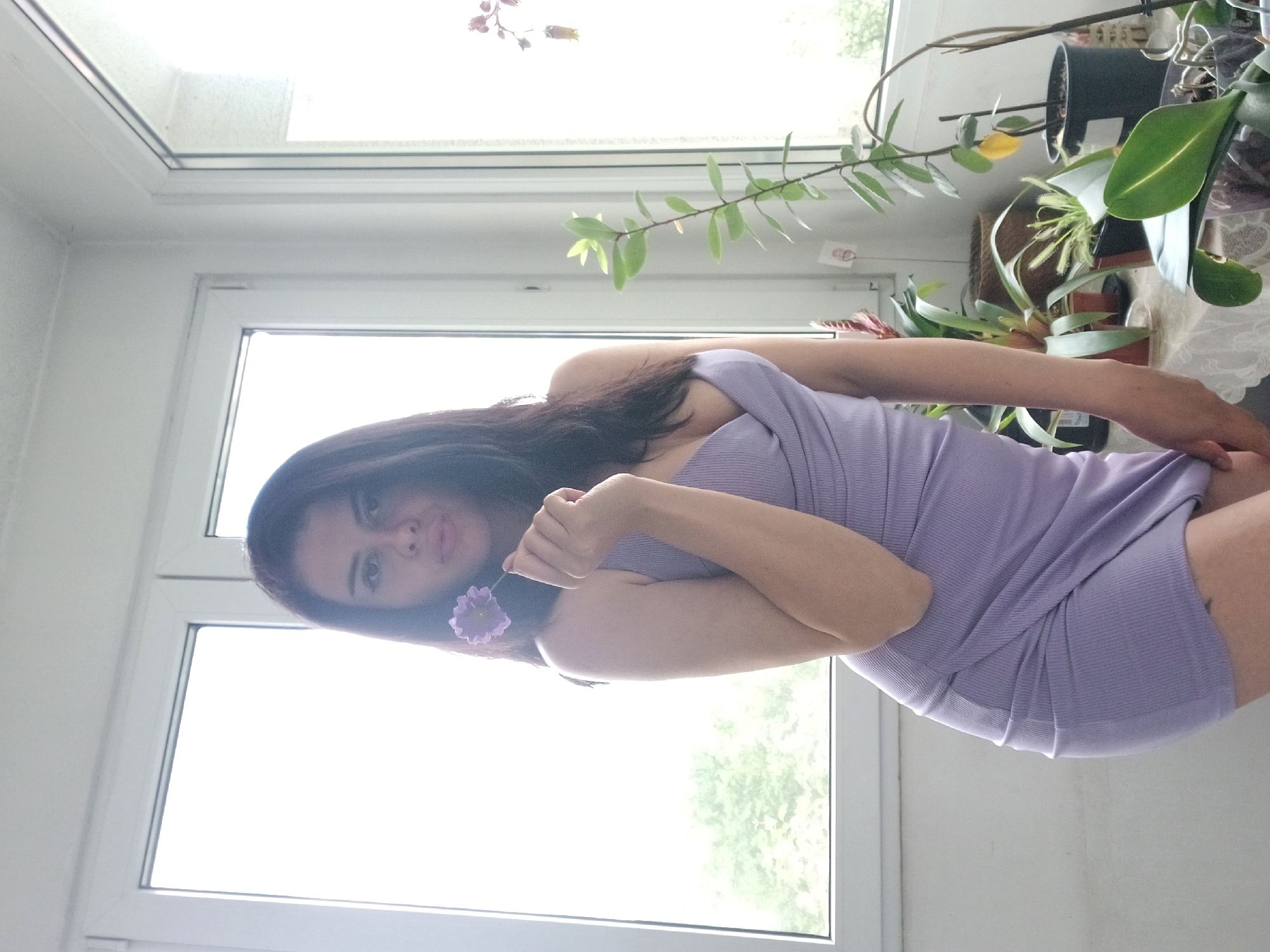 A woman with dark hair is holding a small purple flower next to her face. She is wearing a lavender ribbed dress. The background features a bright window with a view of greenery outside and several potted plants on the windowsill. It is not possible to identify the precise location in Sofia, Bulgaria.