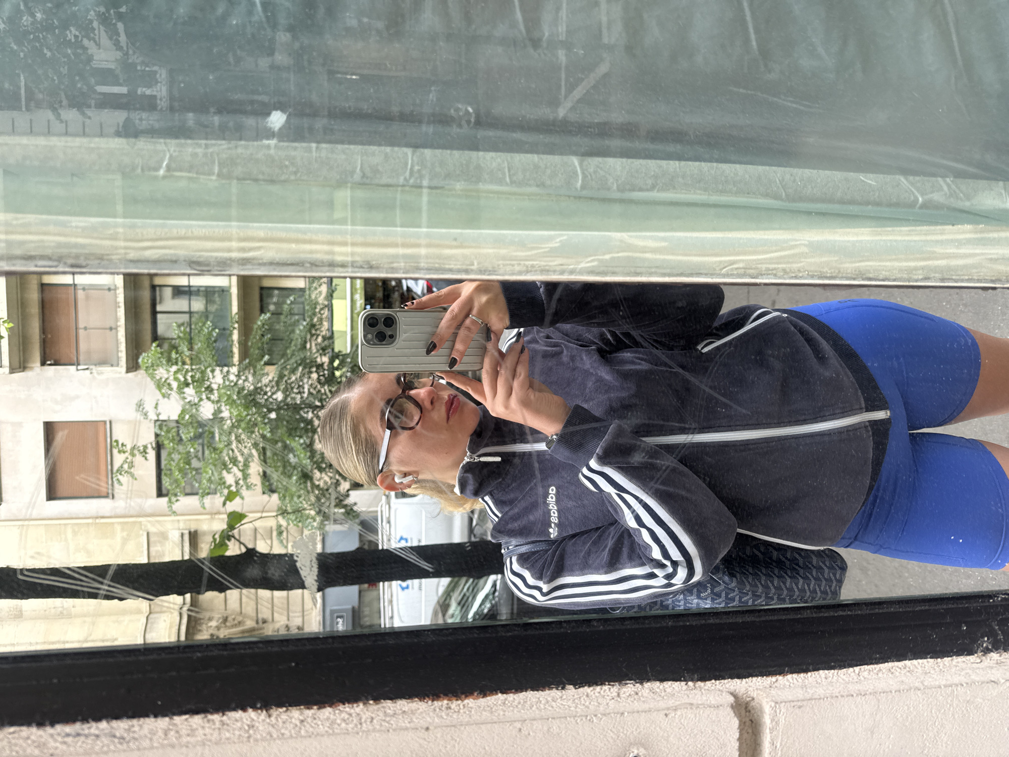 A woman in Paris, France takes a mirror selfie. The woman is wearing blue spandex bike shorts and an Adidas three-stripe track jacket. She is holding a smartphone to take a photo in front of a store window. The window has a reflection of the buildings across the street and a tree. The image also shows a black Goyard handbag next to the woman.