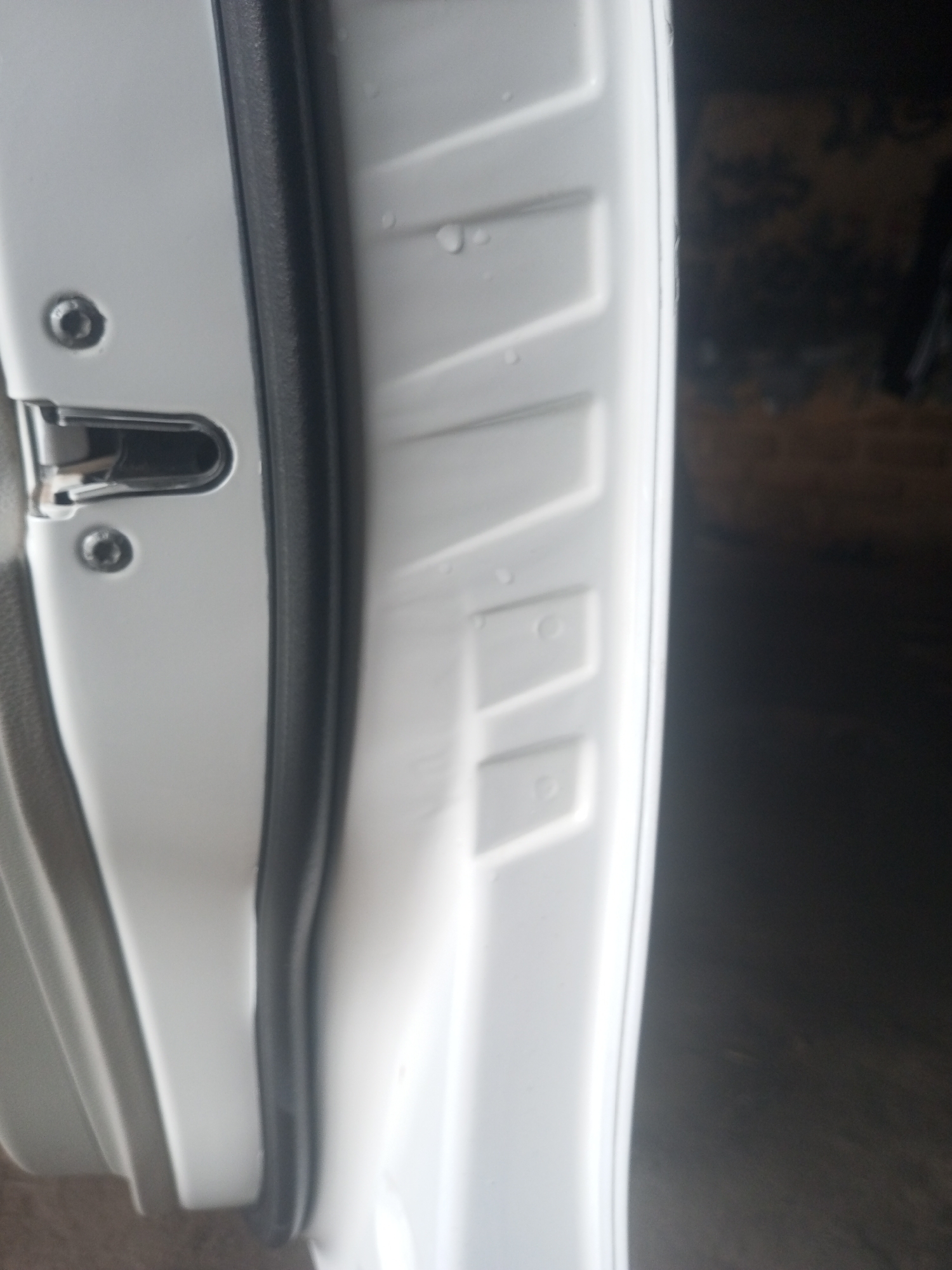 The image shows the interior of a white car door. A black rubber seal runs along the edge of the door. A chrome latch mechanism is visible, as is a circular screw or bolt head. The door panel itself has a textured, ridged surface with some small water droplets. The background is dark and out of focus, suggesting an indoor or shaded environment. The scene is likely within the city of New Stadium, Nigeria, as per the prompt's context.