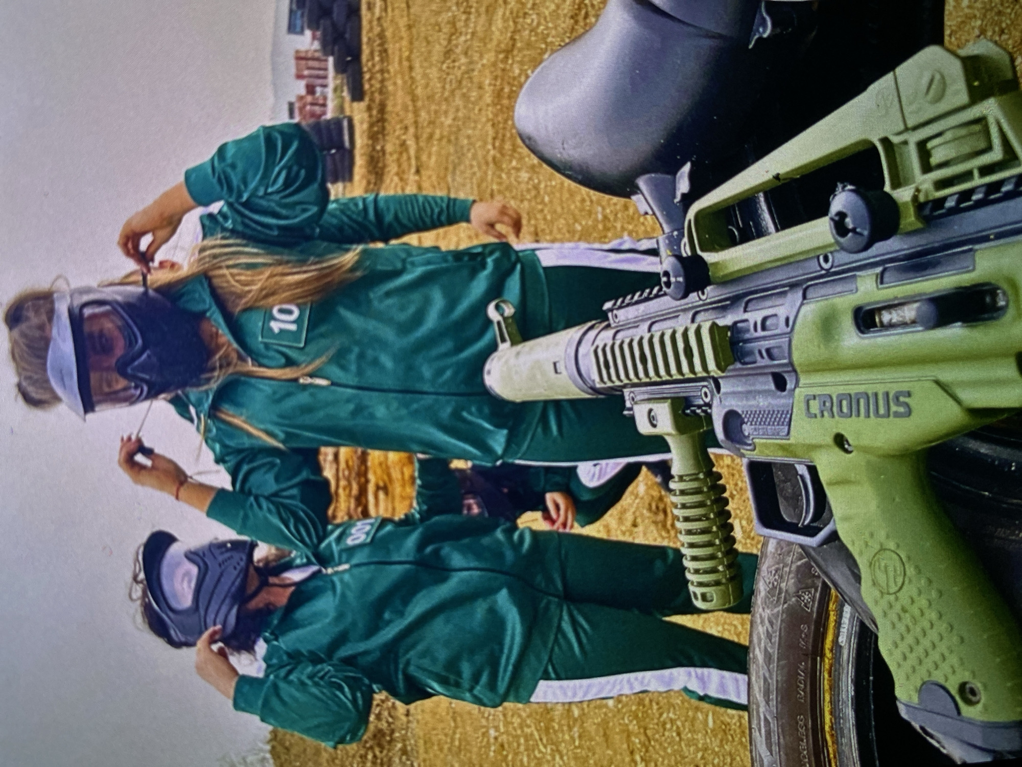 Two individuals are depicted in an outdoor setting, likely a paintball arena in Bankya, Bulgaria. Both are wearing dark green jumpsuits with white side stripes, reminiscent of "Squid Game" player uniforms, and full-face paintball masks for protection.
The person in the foreground on the left has the number "10" visible on their chest and is adjusting their mask or hair. The second person, further back on the right, has the number "08" partially visible on their uniform and is also adjusting something near their face.
A prominent olive green paintball marker is in the foreground, partially obscuring the person on the right. The text "CRONUS" is clearly visible on the side of the marker, identifying it as a Tippmann Cronus model. It features a top-mounted hopper for paintballs and a textured grip.
The ground is dry and earthy, suggesting a dirt or sand field, common for paintball courses. In the background, stacks of black objects, likely tires, serve as barriers or cover. The sky appears bright, indicating daytime. A section of a black tire with text like "TUBELESS RADIAL M+S" is visible beneath the paintball marker in the extreme foreground.