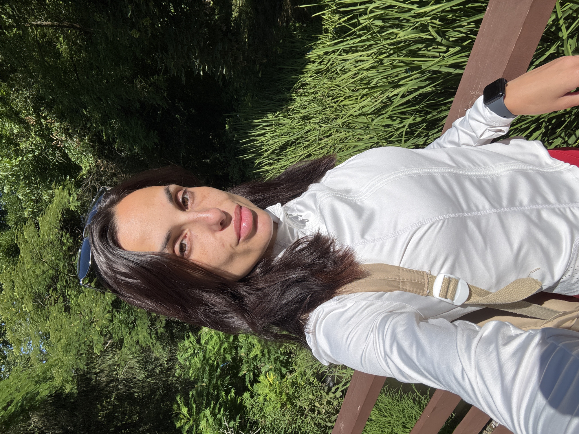 A medium shot shows a woman with dark brown hair and light skin, wearing a white jacket, and a beige backpack strap is visible on her shoulder. She is sitting on a wooden bench in an outdoor setting with lush green trees and tall grass in the background. The sun is shining brightly, casting shadows. The woman is looking towards the camera, with sunglasses resting on top of her head. Her smartwatch is visible on her left wrist. The scene appears to be a park or garden in Pleven, Bulgaria.