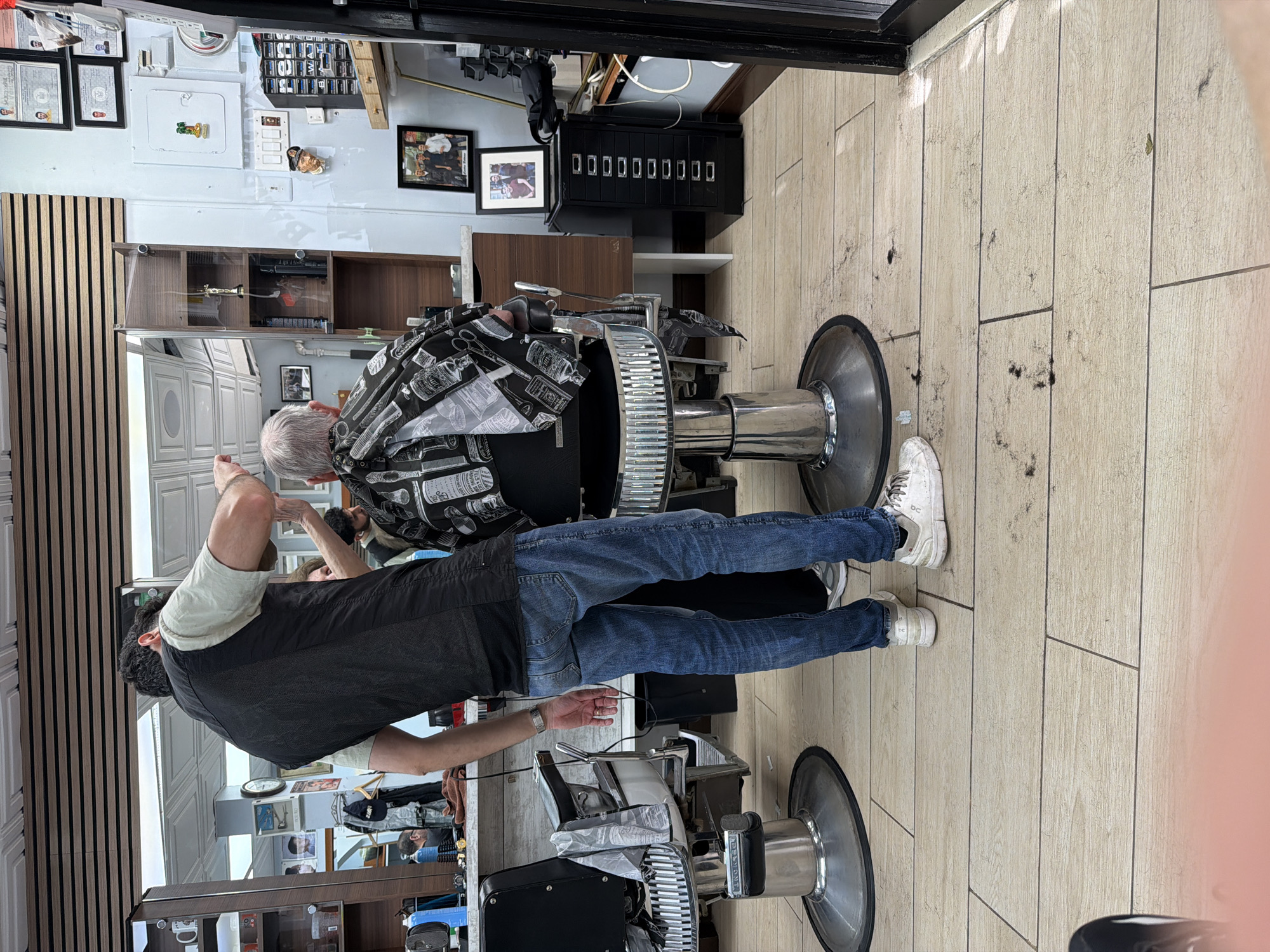 A barber gives a haircut to a man in a barbershop. The barber is wearing a black vest over a white shirt and blue jeans and has his back to the camera. He is cutting the hair of an older man who is sitting in a barber chair. The floor is covered in hair. The barbershop is located in New York City, United States.