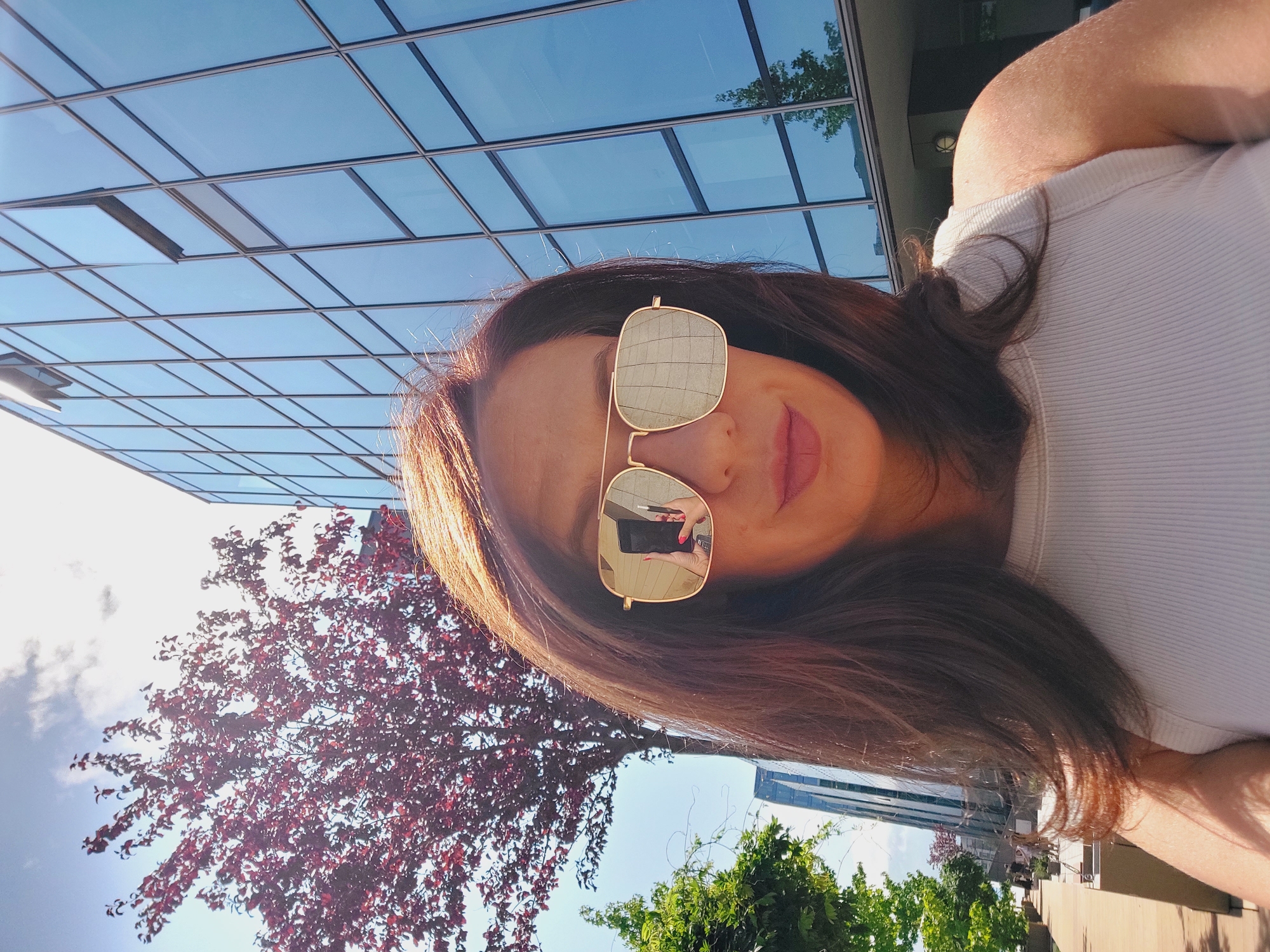 In a selfie taken outdoors, a woman with shoulder-length brown hair is featured. She is wearing large, gold-rimmed aviator sunglasses with reflective lenses. Her mouth is slightly parted, and her lips are painted with a subtle pink lipstick. She is wearing a white, ribbed tank top. In the reflection of her sunglasses, a smartphone can be seen being held by a hand with red nail polish, and a stylus or pen.
The background suggests an urban environment. To the left, a tree with dark red leaves is visible against a bright blue sky with scattered white clouds. Further left, a smaller green tree and a modern glass-fronted building are present. To the right of the woman, a large, multi-story building with a facade of blue glass windows is dominant. The windows reflect the sky and surrounding trees. The overall lighting indicates it is a sunny day. The image was taken on the pedestrian zone in front of the National Palace of Culture in Sofia, Bulgaria, indicated by the distinct architecture of the building and the style of the surrounding area.