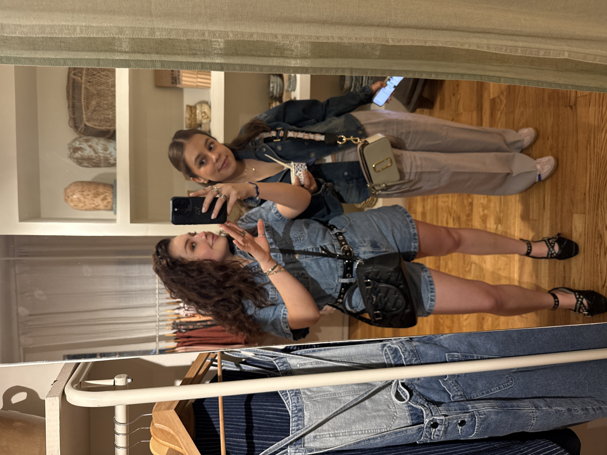 Two young women are reflected in a mirror inside a retail store, potentially in Mexico City, Mexico. One woman is wearing a denim jumpsuit, a black belt, and black sandals, and she is holding a smartphone. The other woman is wearing a blue denim jacket, a white top, tan pants, and light-colored shoes, and she is holding a smartphone. The mirror reflects shelves with baskets and decorative items, as well as clothing on racks. The floor is made of wood planks.