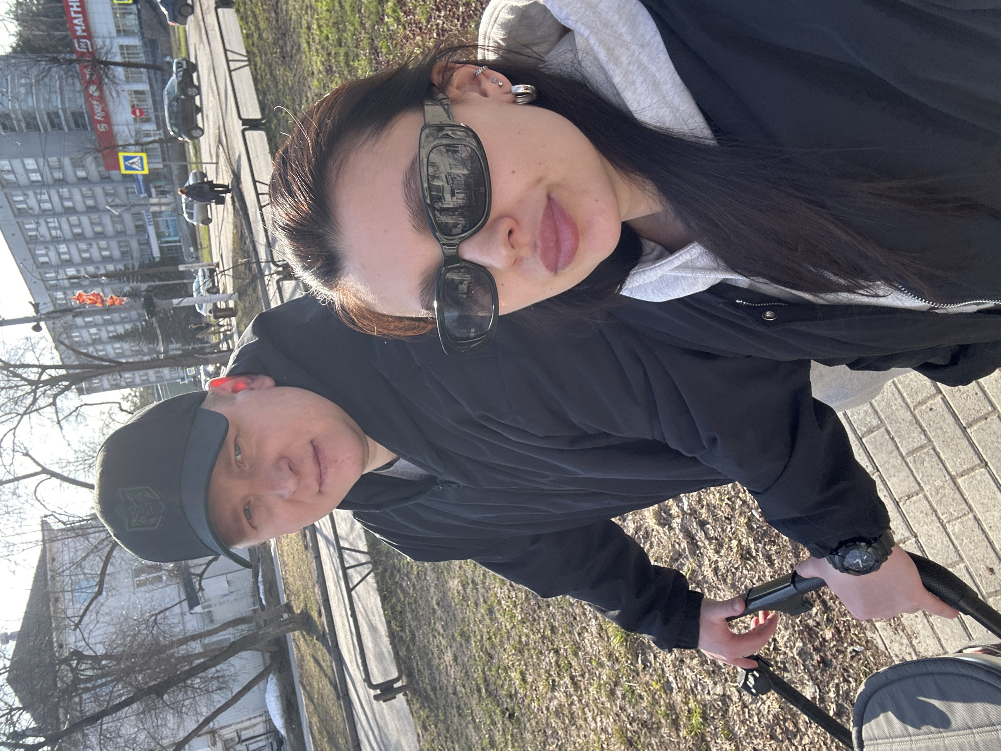 Two people are taking a selfie on a street in Chaikovsky, Russia. The woman is in the foreground, wearing sunglasses, a gray hoodie, and a black jacket. The man is behind her and to the left, wearing a black baseball cap with the coat of arms of Ukraine. They are on a paved area next to a grassy area with trees, possibly pushing a stroller. In the background, there's a multistory building and cars on a road. A pedestrian crossing sign is also visible.