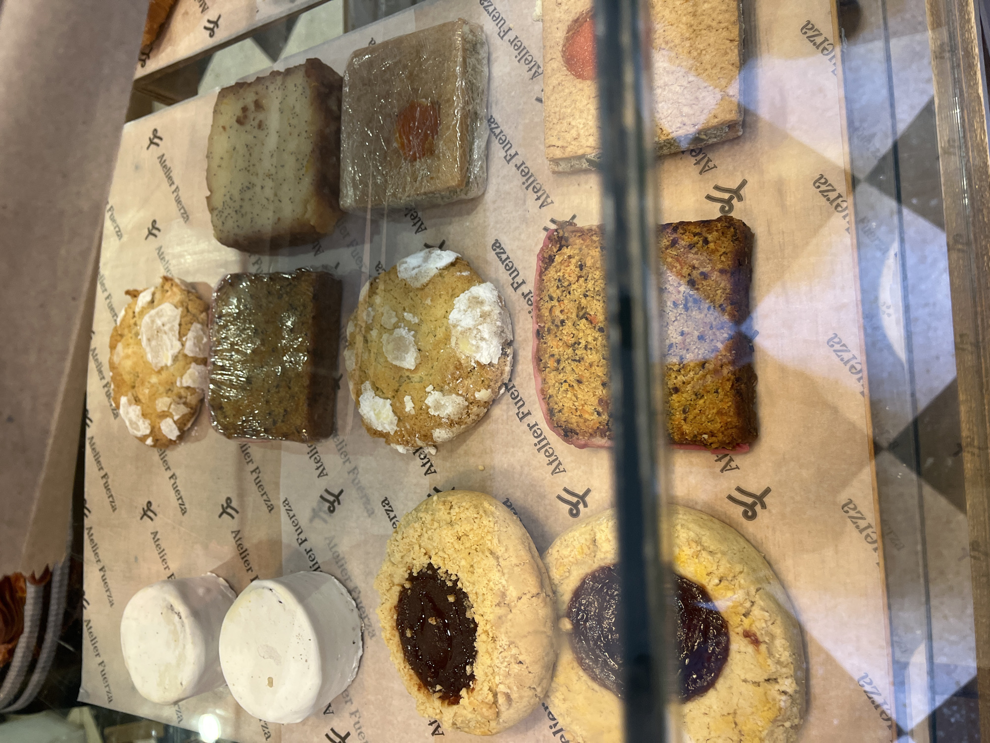 This close-up shot captures a variety of baked goods and pastries displayed in what appears to be a bakery or cafe in Olivos, Argentina. The items are arranged on light brown parchment paper, visible through a glass display case with reflections.
On the top row, from left to right, there is a rectangular loaf or cake speckled with poppy seeds, followed by a square pastry wrapped in plastic, featuring an orange-colored fruit piece on top. Another light-colored rectangular item is partially visible to the far right.
The middle row showcases a round, light-colored cookie or scone with white frosting or sugar, next to a darker rectangular item wrapped in plastic. Another similar round cookie/scone is to its right, and then a reddish-brown, textured rectangular cake, possibly a carrot or fruit cake.
The bottom left of the display holds two small, cylindrical, white items. To their right are two large, round, light-colored cookies with a dark, jam-like fruit filling in their centers.
No people are visible in the image. The parchment paper beneath the items is adorned with a repeating logo and the text "Atelier Fuerza," which is also fragmented into "erza," "A," "elier," and "Fuer" across the different visible sections. The logo appears to be a stylized 'AF' or two overlapping 'f's. The reflections on the glass and a vertical metal frame indicate the items are behind a transparent barrier. The time of day and weather are not discernible.