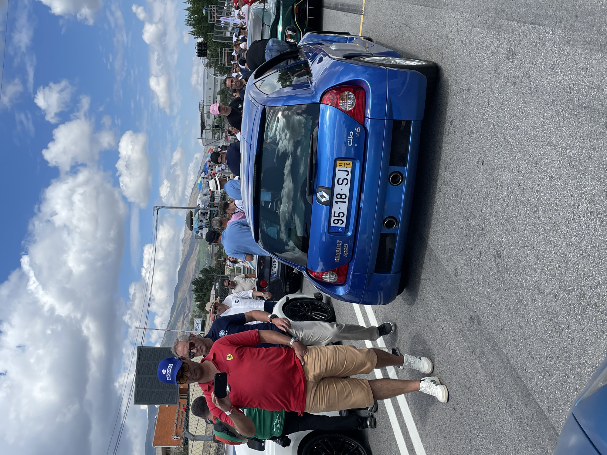 This image depicts an outdoor event, likely a car show or race, at the Circuito de Vila Real in Portugal. The scene is crowded with people and several vehicles are visible. In the foreground, a bright blue Renault Clio V6 is parked on an asphalt surface marked with white lines. The car has a license plate reading "95-18-SJ".
To the left of the Clio, a man wearing a red polo shirt with a yellow Ferrari logo and a blue baseball cap is holding up a smartphone, appearing to take a photo or video. He is wearing tan shorts and white sneakers. Behind him, other attendees are visible, some looking towards the cars or interacting with each other. A white car is partially visible to the left of the man in the red shirt.
In the background, more cars, including a dark grey Corvette and a green sports car, are present. A crowd of people stands around the vehicles, and tents and structures are set up in the distance. The environment includes hills with vegetation and buildings in the far background, under a sky with scattered white clouds. The overall atmosphere suggests a sunny day at an automotive event.