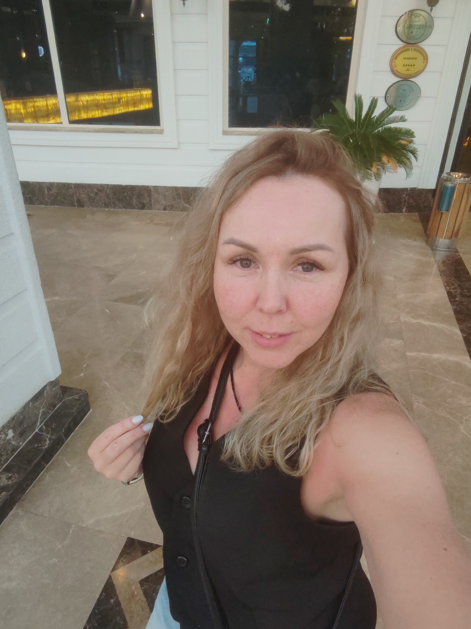 A woman with fair skin and blonde, wavy hair is posing for a selfie. She has freckles on her face and is wearing a black top with buttons. She is standing indoors on a patterned marble floor. In the background, there are white columns, windows, and a potted palm tree. On the wall, there are decorative medallions. Based on the decor and the presence of what appears to be hotel awards/decorations, the location is likely a hotel lobby or upscale interior in Alanya, Turkey.