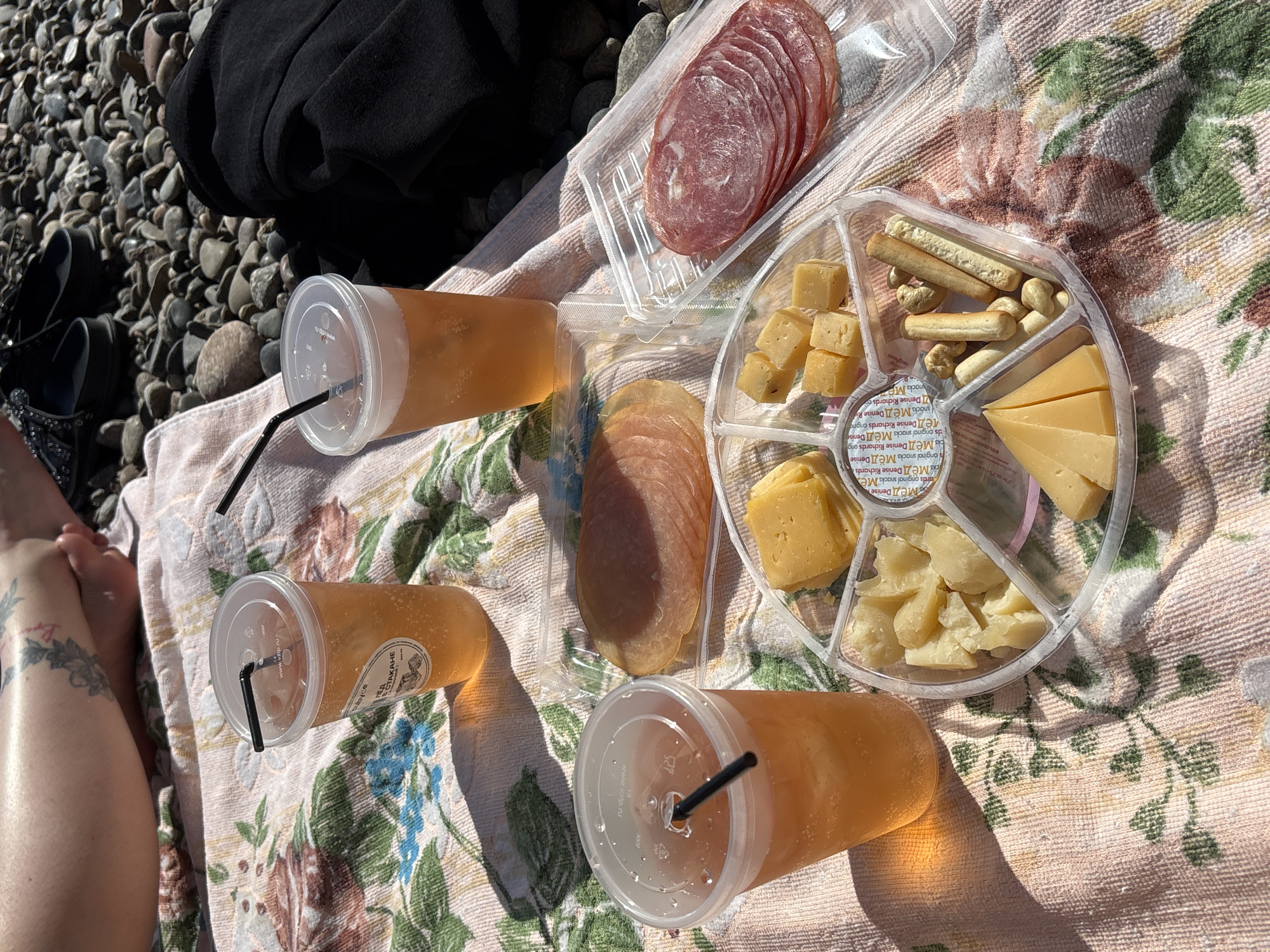 This medium shot captures a sunny outdoor picnic scene, likely by a body of water given the extensive pebbled ground visible to the left. A person's legs and feet, one adorned with a tattoo, are visible on the left, resting on a floral-patterned blanket.
The main focus is a spread of food and drinks arranged on the blanket. Three clear plastic cups with black straws hold an amber-colored beverage, possibly iced tea or a light juice. Two plastic containers of sliced deli meats, including salami and another lighter-colored cured meat, are present. A circular, multi-compartment plastic platter features a variety of cheeses (cubes, wedges, and irregular pieces), breadsticks, and nuts (likely cashews).
The scene suggests a relaxed daytime gathering. Bright sunlight casts distinct shadows, indicating a clear, sunny day. The pebbled ground implies a natural setting such as a beach, riverbank, or lakeside shore, consistent with outdoor leisure in a location like Krasnoyarsk, Russia.
Visible text includes "www.vzlp.ru" on the lids of the cups. Labels on the small packages within the cheese platter read "МЁД Denise Richards original snacks" and variations thereof (МЁД means "Honey" in Russian). One cup has a label with "В СТАКАНЕ" (V STAKANE, meaning "In the cup") and "МЁД" (Honey), while another label reads "cubyce".