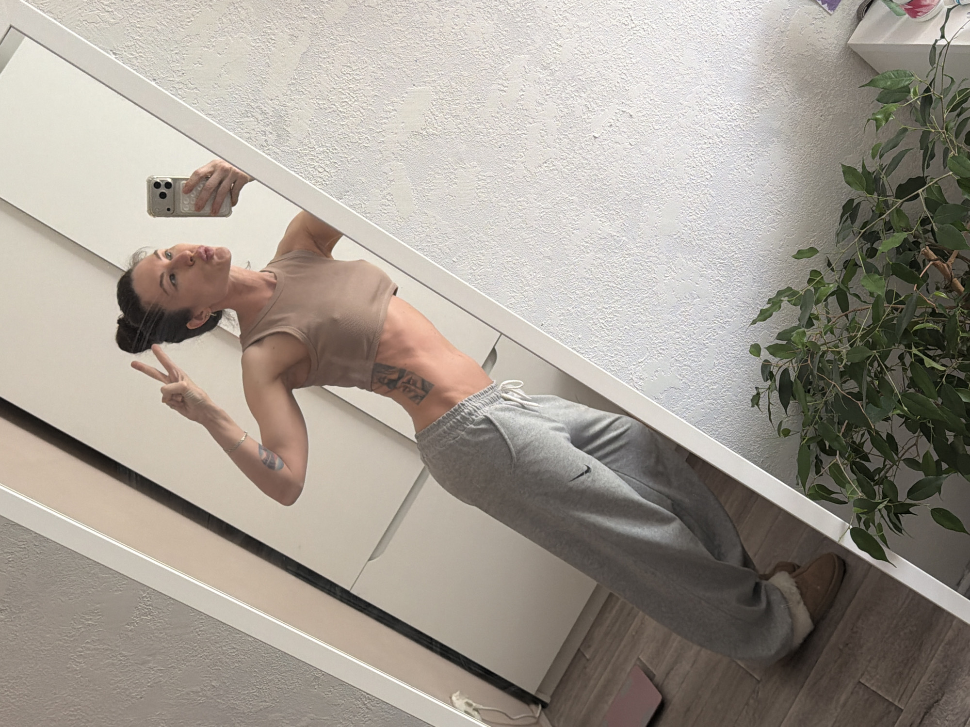 A woman is taking a selfie in a mirror. She is wearing a tan crop top and gray sweatpants. She is holding up a peace sign with her left hand. The background includes a white wall, a white wardrobe, a potted plant, and a wooden floor. There is no information to suggest that the location is Perm, Russia.