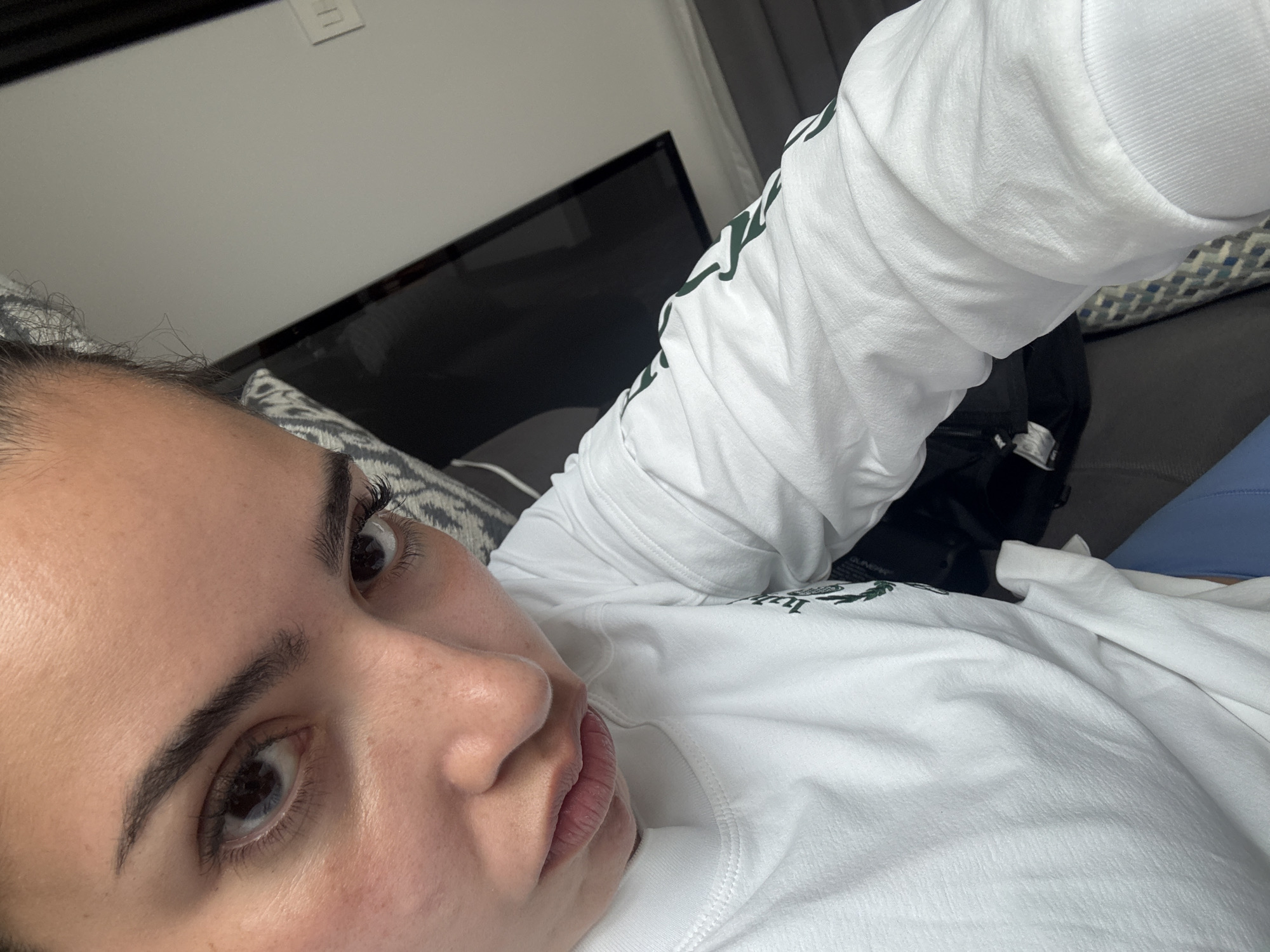 A selfie shot from a low angle shows a person with fair skin, dark eyebrows, and brown eyes. The person is wearing a white long-sleeved shirt with dark green detailing on the sleeve. The person appears to be lying down on a couch with a gray and white patterned pillow behind them. In the background, there is a black television screen and a light gray wall with a light switch. A portion of a dark gray curtain is visible on the right side of the frame. There is no evidence of the location being San Bernardino Tlaxcalancingo, Mexico.