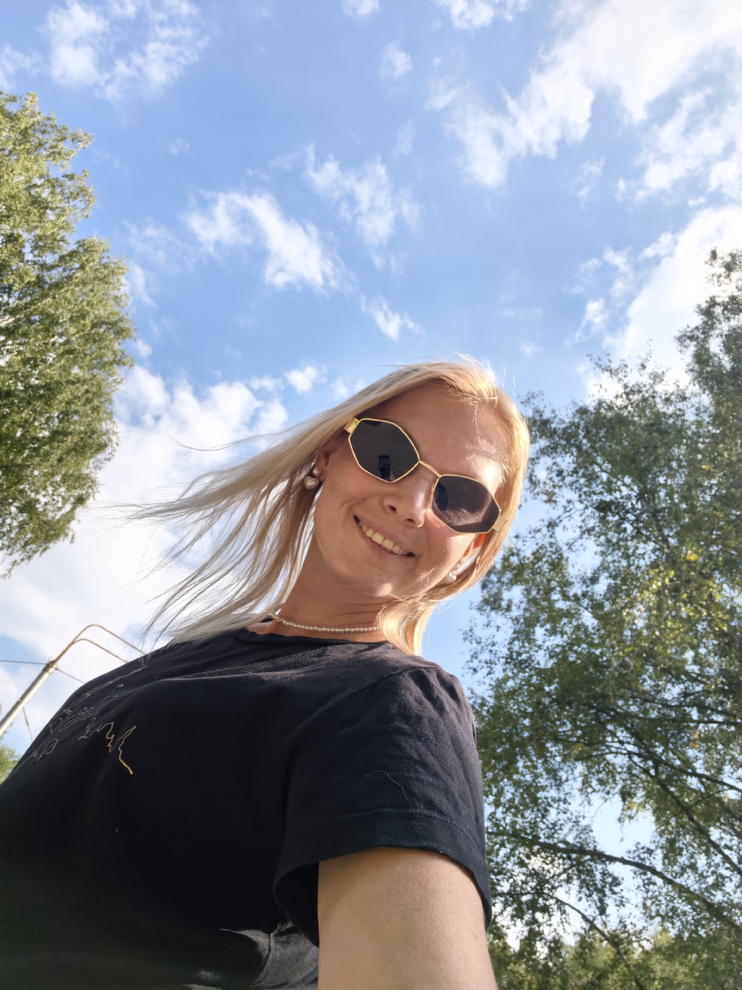 A young woman with blonde hair, wearing sunglasses and a black t-shirt, is pictured from a low angle against a blue sky with white clouds. She is smiling and her hair is blowing in the wind. Green trees are visible on either side of the frame, suggesting an outdoor setting. The image was captured in Sterlitamak, Russia.