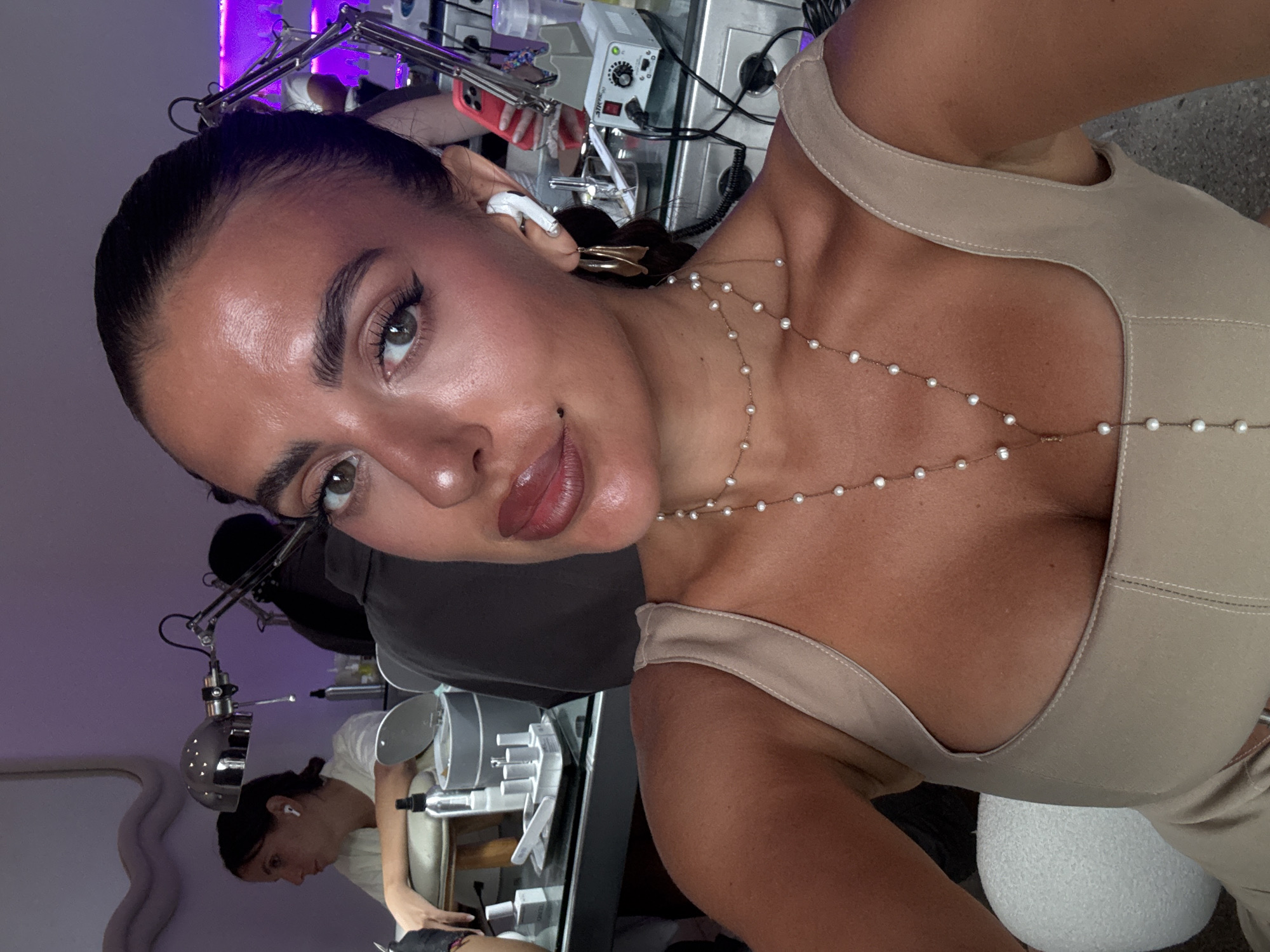 The image is a close-up selfie of a woman with dark hair pulled back, wearing a beige top and a pearl body chain. She has winged eyeliner and glossy lips. Behind her, partially visible, is a beauty salon setting with equipment, including a lamp and various machines. Another person, presumably a beautician, is visible in the background working on a client. The presence of equipment and the beautician suggest that this image was taken in a beauty service establishment, likely offering treatments like brow shaping or cosmetic tattooing. The woman in the foreground is looking directly at the camera, possibly capturing a moment before or after a beauty treatment. The location is possibly North Kuta, Indonesia.