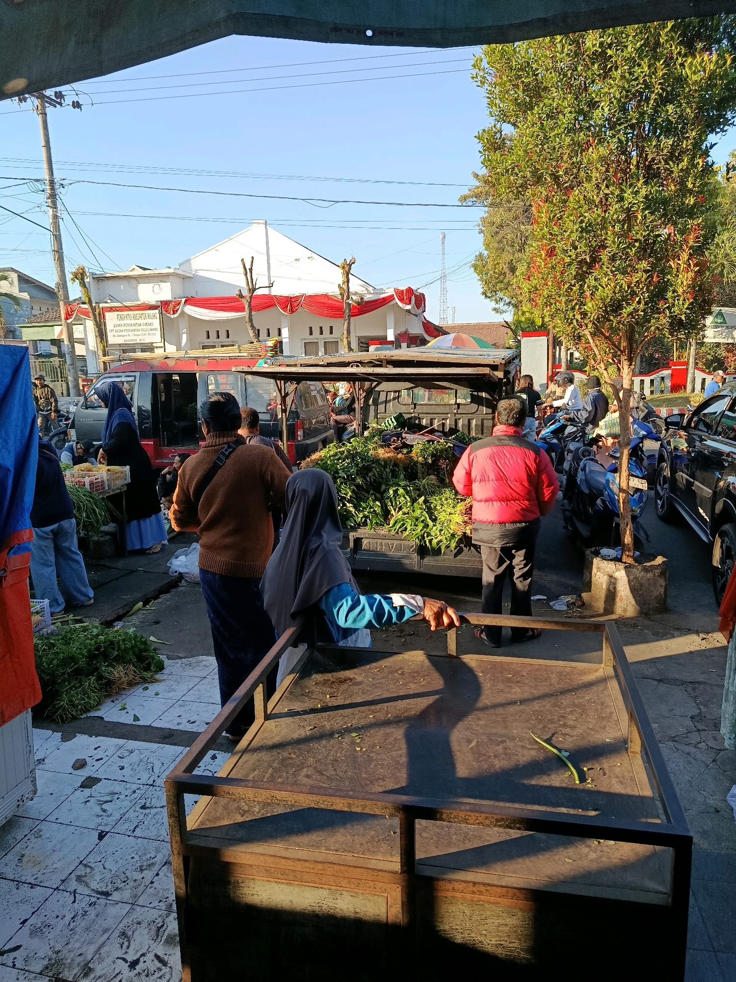 A street market scene unfolds in Bululawang, Indonesia, under a clear blue sky. The foreground features a weathered, dark brown metal cart with an empty top surface, casting long shadows. Behind it, a variety of people are engaged in commerce. To the left, a woman wearing a blue hijab and white dress stands near a red van, attending to a basket of produce. A man in a brown sweater and dark pants is positioned centrally, facing away from the camera, with a black strap across his shoulder. To his right, a person in a red jacket and black pants examines a large pile of green vegetables on the back of a dark colored truck. Further back, several motorcycles are parked, and a dark blue SUV is visible on the right side of the frame. The background shows a white building adorned with red and white festive banners, suggesting a possible local event or government facility. A tall tree with green foliage stands to the right, adding to the outdoor market ambiance. The ground is paved with light-colored tiles, some of which are worn and stained. Electrical poles and wires are present on the left, indicating an urban setting.