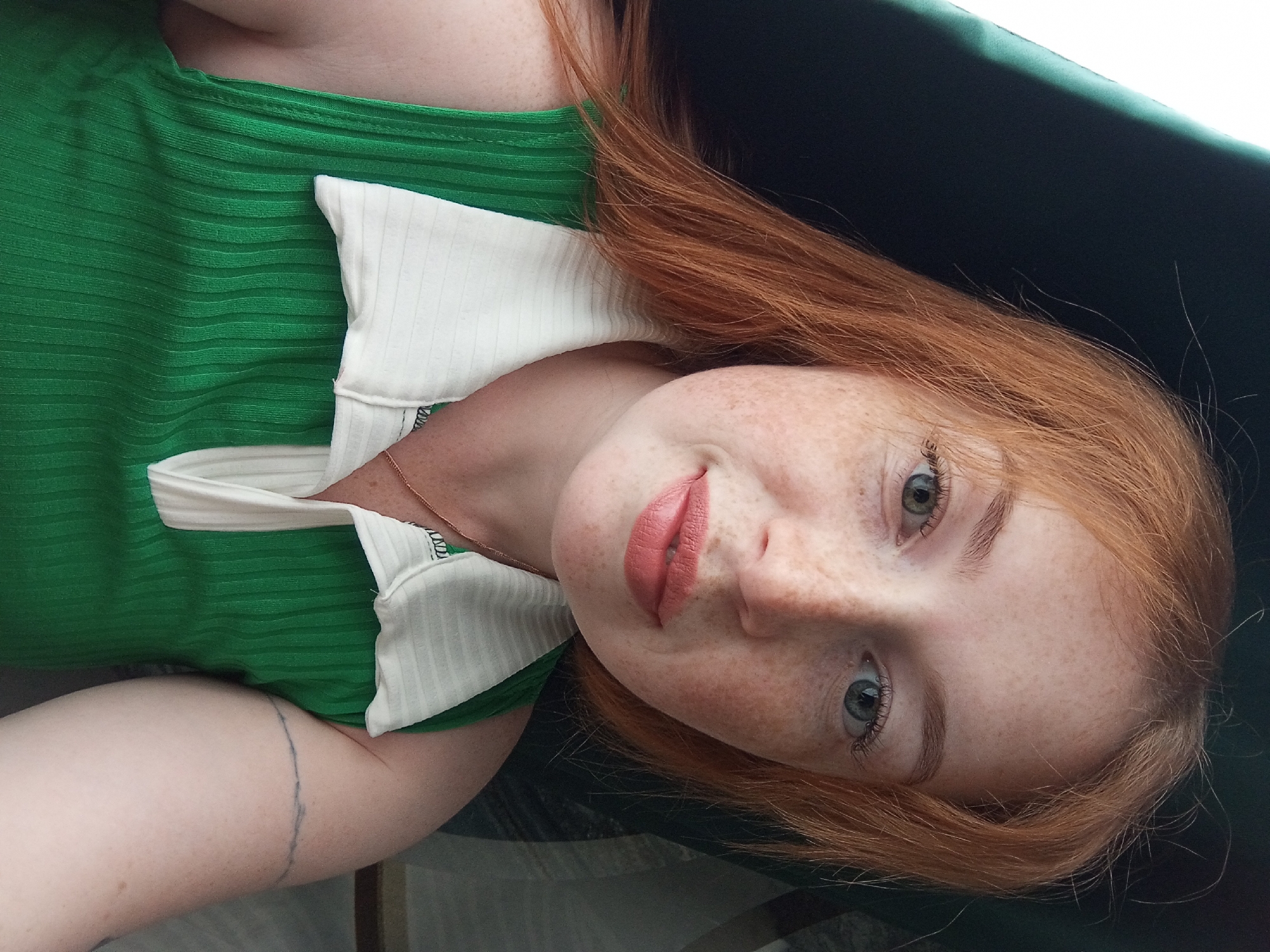A young woman with auburn hair and freckles is lying down, looking up at the camera. She is wearing a green ribbed top with a white collar. A dark green surface is visible behind her head. A tattoo is visible on her left upper arm. The location cannot be determined from the provided image.