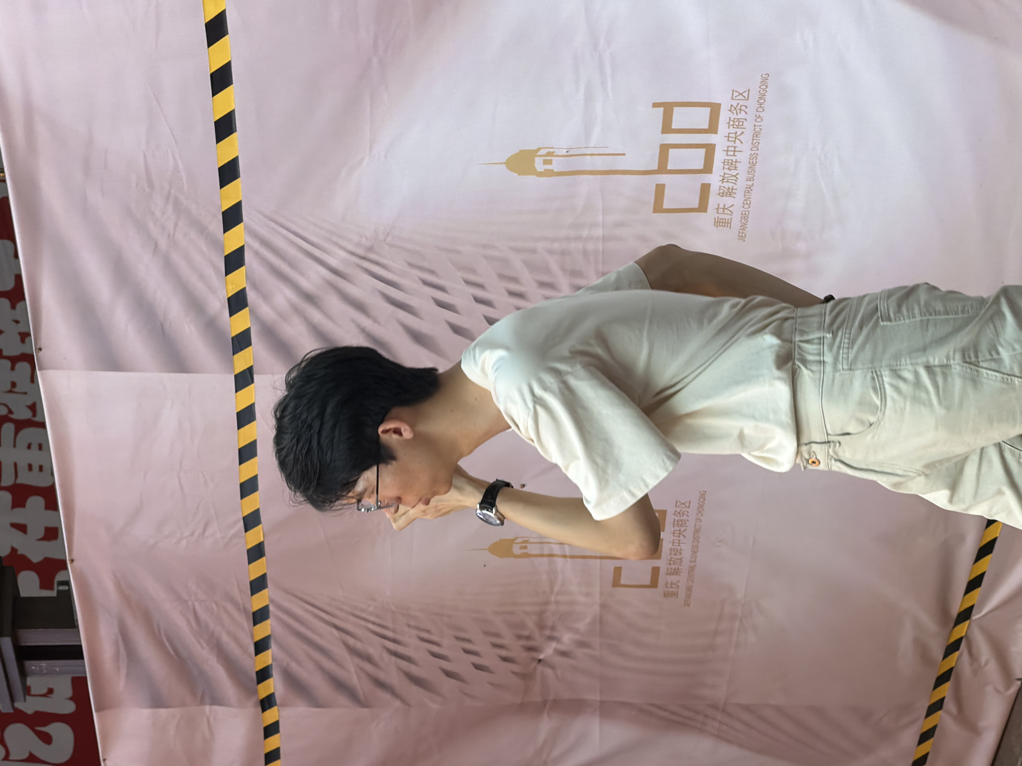 A young Asian person with dark hair and glasses is bent over, touching their forehead to a large, light pink banner. They are wearing a light-colored, short-sleeved t-shirt and light-colored pants. A black and yellow striped ribbon is visible on the left side of the banner. The banner features gold text and a stylized graphic of buildings. The text on the banner reads "重庆解放碑中央商务区 JIEFANGBEI CENTRAL BUSINESS DISTRICT OF CHONGQING". The person appears to be in a public space, possibly an outdoor event or exhibition given the banner. The lighting suggests it is daytime.