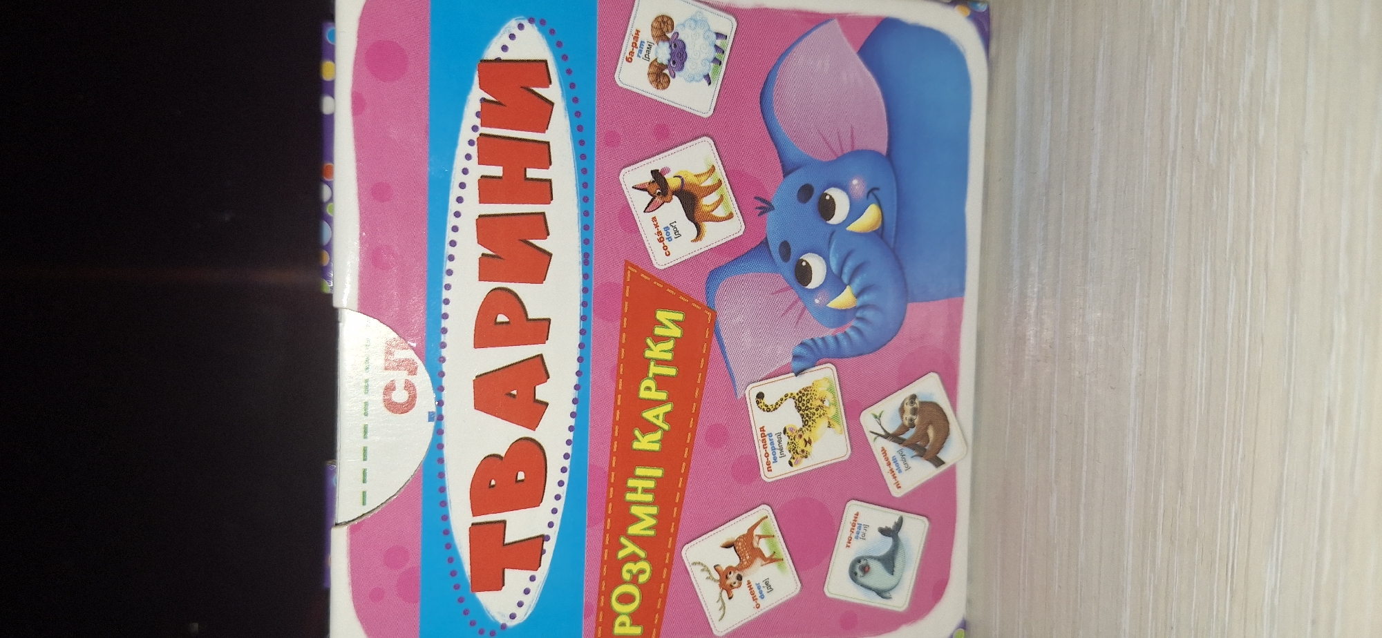 The image shows the front of a children's game box titled "ТВАРИНИ" (Animals) in large red letters. Below this, "РОЗУМНІ КАРТКИ" (Smart Cards) is written on an orange banner. The box has a pink background with white polka dots and a blue border with purple dots. Several cards with cartoon animals are displayed around a blue elephant illustration. These include a deer, a leopard, a sloth, a seal, a dog, and a ram. The box appears to be new and is placed on a light-colored wooden surface. There is no visible context that would indicate its location in Bila Tserkva, Ukraine.