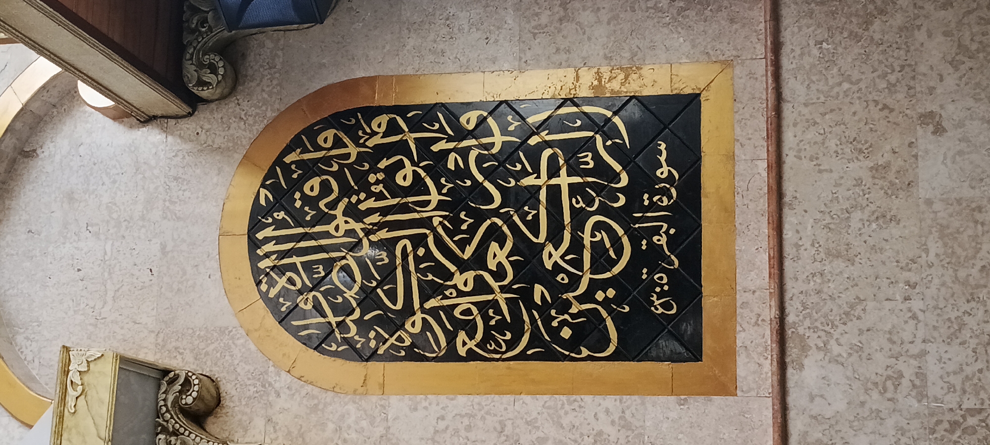 The image shows a close-up of an arched wooden panel with Islamic calligraphy in gold lettering on a black background. The panel is framed by a golden border and is set into a textured, light-colored wall. The calligraphy appears to be verses from the Quran, as indicated by the text at the bottom of the panel which reads "سورة البقرة ٤٣:٥" (Surah Al-Baqarah 43:5). The wall is made of rectangular tiles with a speckled pattern. There are decorative architectural elements visible in the upper left and upper right corners of the image, suggesting this is an interior or covered space within a building. The overall impression is one of religious decoration within a building, possibly a mosque or a prayer room, located in Denpasar, Indonesia. The image is captured from a slightly upward angle.