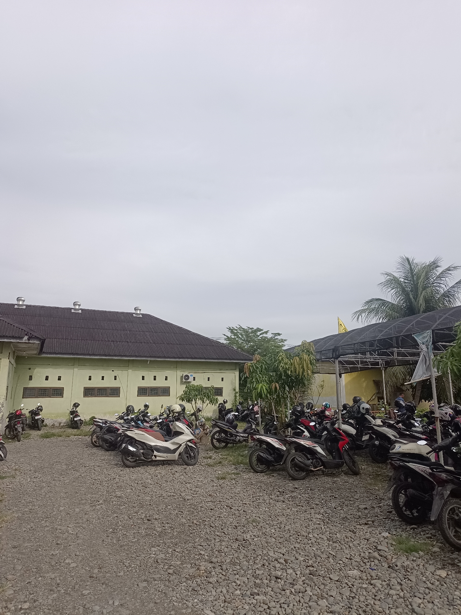 The image depicts an outdoor scene with a building and a parking area filled with motorcycles. The building is light green with a dark, corrugated metal roof. It has rows of rectangular windows and several circular vents on the roof. To the right of the building, there is a covered structure with a black mesh roof supported by poles. A yellow flag is visible on a pole behind this structure. Palm trees and other foliage are present in the background. The foreground is covered in gravel and grass, serving as a parking lot for numerous motorcycles, most of which are parked and unoccupied. Several motorcycles are arranged in rows, with helmets placed on some of them. A few individuals can be seen in the background, near the covered structure. The sky is overcast and grayish. The location is identified as Mesjid Reuntoh, Indonesia.