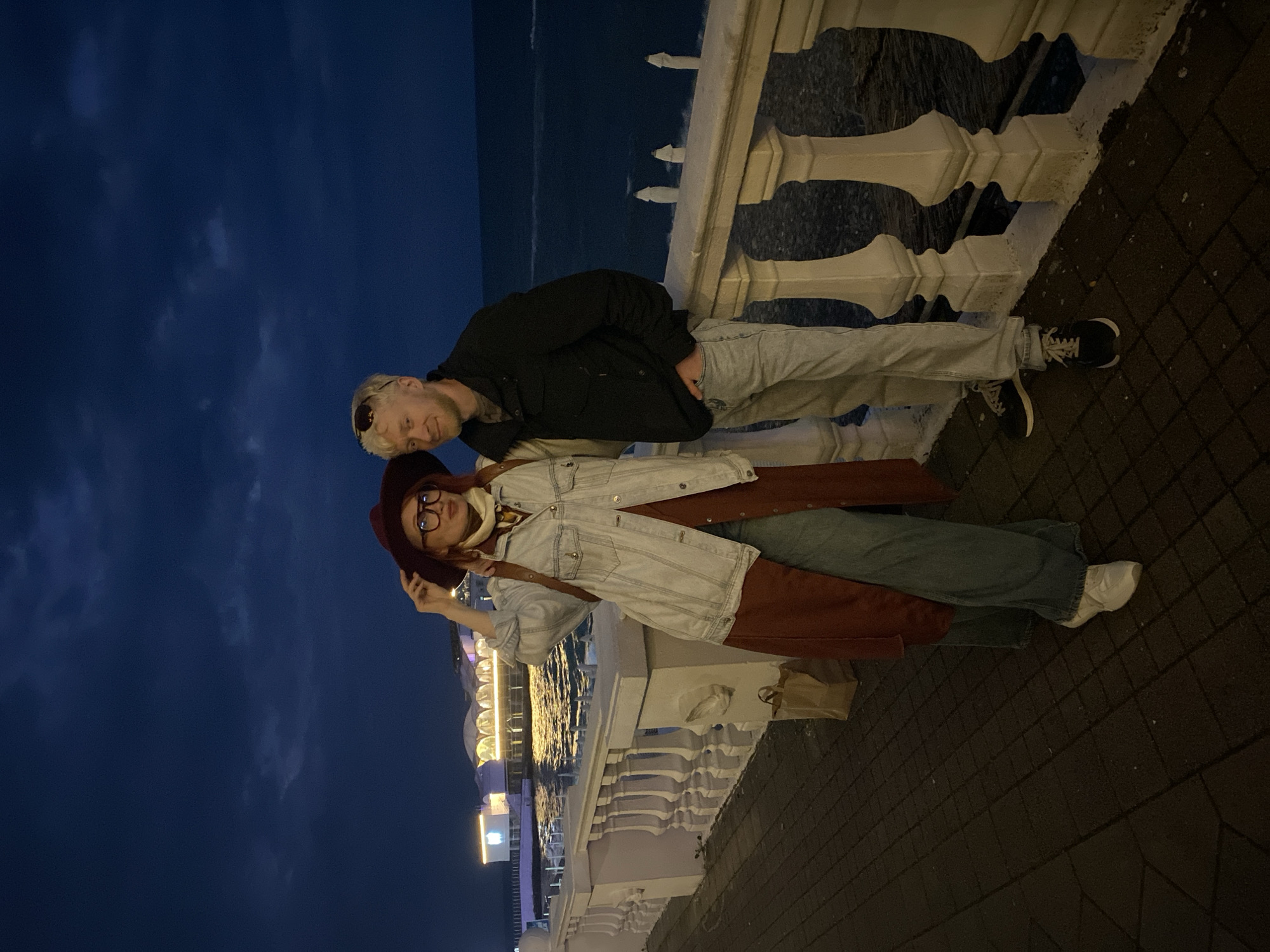 Two people are posing for a photograph outdoors at night. The woman, on the right, is wearing a maroon hat, eyeglasses, a white scarf, a light-colored denim jacket, a rust-colored garment underneath, and blue jeans. She is holding her hat. The man, on the left, is wearing a dark jacket and light-colored pants. They are standing next to a white decorative railing. Behind the railing is a body of water, and in the distance, there are lighted buildings. The background sky is dark. The patterned ground suggests they are on a walkway. The lighted buildings in the distance suggest the architecture and illuminated structures are characteristic of Sochi, Russia.