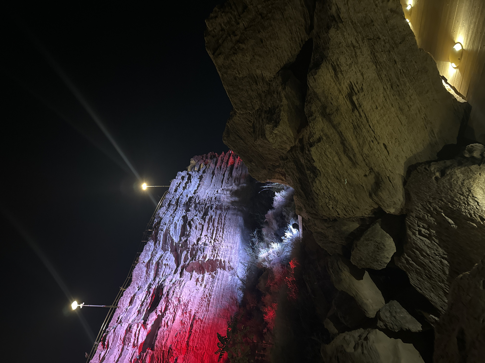 This is a nighttime photograph of a rocky cliff face illuminated by red and white lights. Two lampposts are visible on the top of the cliff, with one light beam extending into the dark sky. Large rocks are in the foreground, and a wooden walkway with small lights is visible at the bottom right. The scene appears to be a scenic overlook or park area, possibly at the Karagiyen Karataba cliff in Aktau, Kazakhstan, known for its unique geological formations and nighttime illumination. There are no people clearly visible in the image. The environment is rugged and natural, with the artificial lighting creating a dramatic contrast with the dark sky.