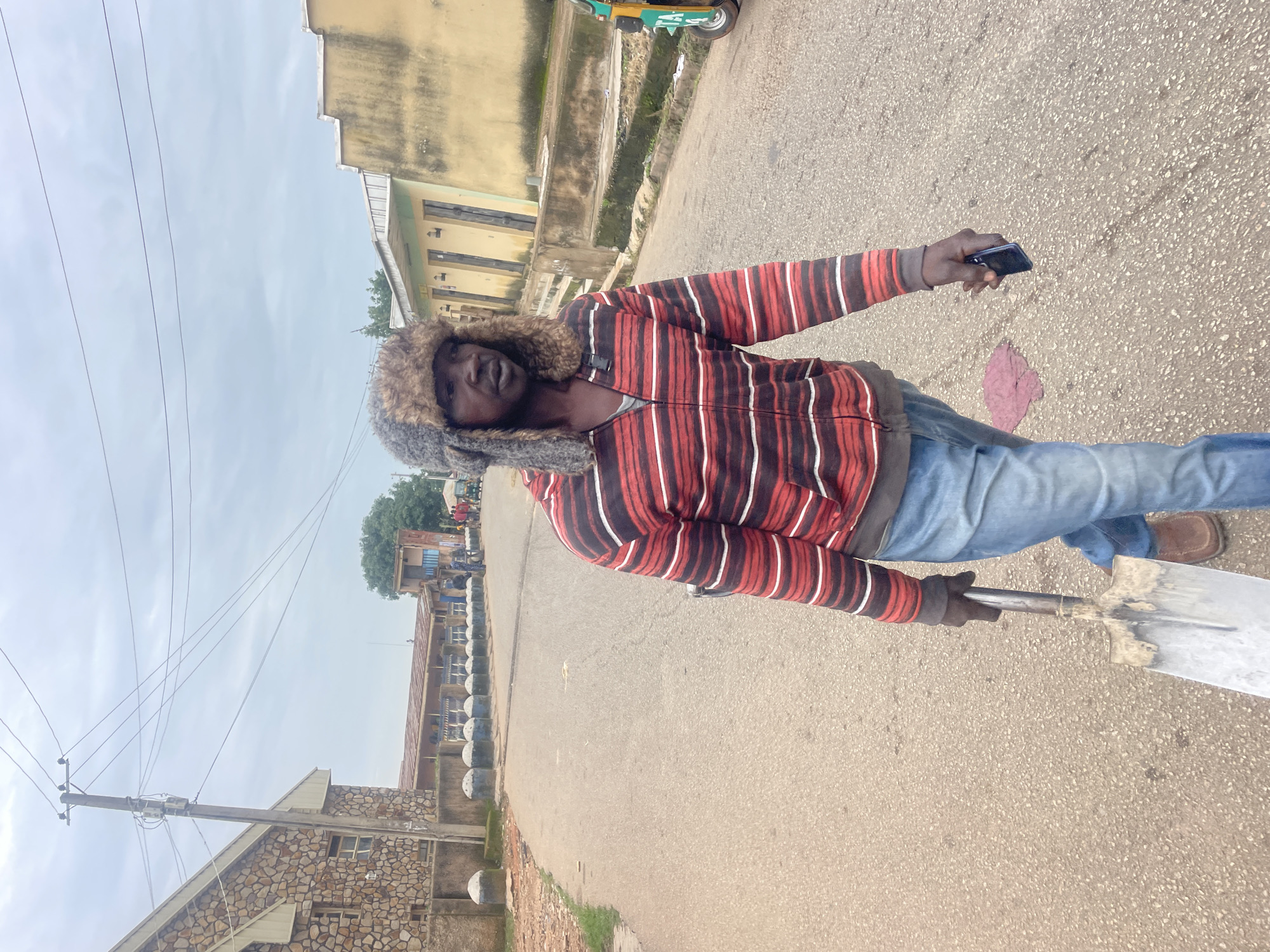 A man is walking down a street in Jos, Nigeria, holding a shovel and a mobile phone. He is wearing a striped hooded sweatshirt, blue jeans, a fur-lined hat, and brown shoes. In the background, there are buildings, a utility pole with wires, and a tricycle. The sky is cloudy. The street is paved with asphalt and appears to be a road in a residential or commercial area. There are some people walking and standing in the distance.