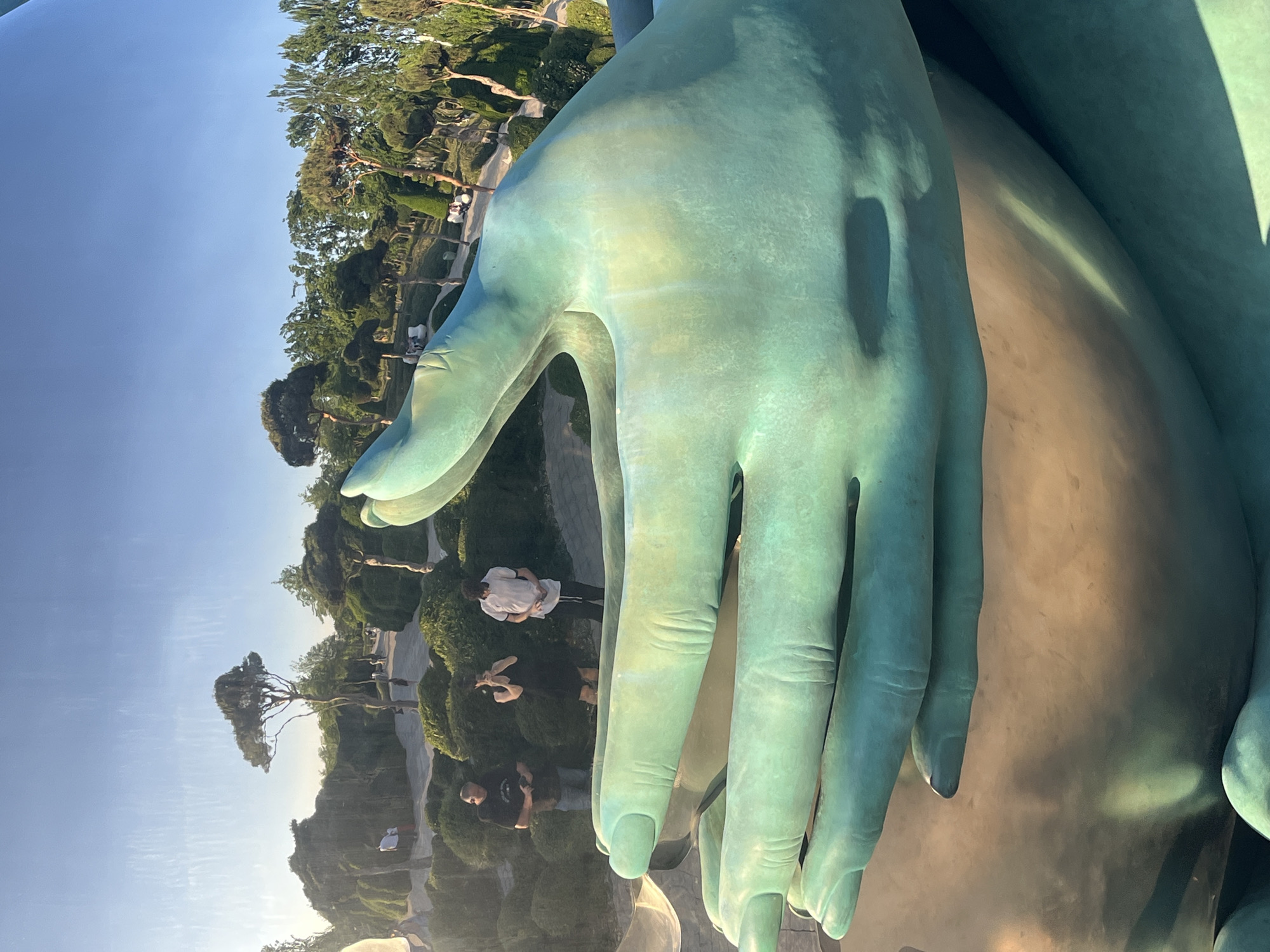 The image displays a colossal, greenish-bronze sculpture of a human hand and part of an arm, dominating the foreground. The statue's surface is highly reflective, acting as a mirror to its surroundings.
Reflected on the glossy surface of the sculpture is a clear, bright blue sky, indicating daytime. Below the sky, numerous trees, manicured bushes, and pathways are visible in the reflections, suggesting the sculpture is situated within a well-maintained park or landscaped garden. Several people are also reflected on the statue's surface. They appear to be walking, standing, and observing the park environment, with some potentially interacting with or photographing the sculpture itself. One person in a white shirt and dark pants is clearly reflected on the palm of the hand, while others are less distinct further back.
The time of day appears to be sunny, given the bright illumination and visible highlights on the sculpture. There is no visible text in the image.
Given the context, this large, reflective art installation is located in Krasnodar, Russia, likely within a public park, possibly the well-known Galitsky Park (Krasnodar Park), which features unique artistic installations of this nature. The overall scene captures an artistic interpretation of a human form integrated with its natural and human environment through reflection.