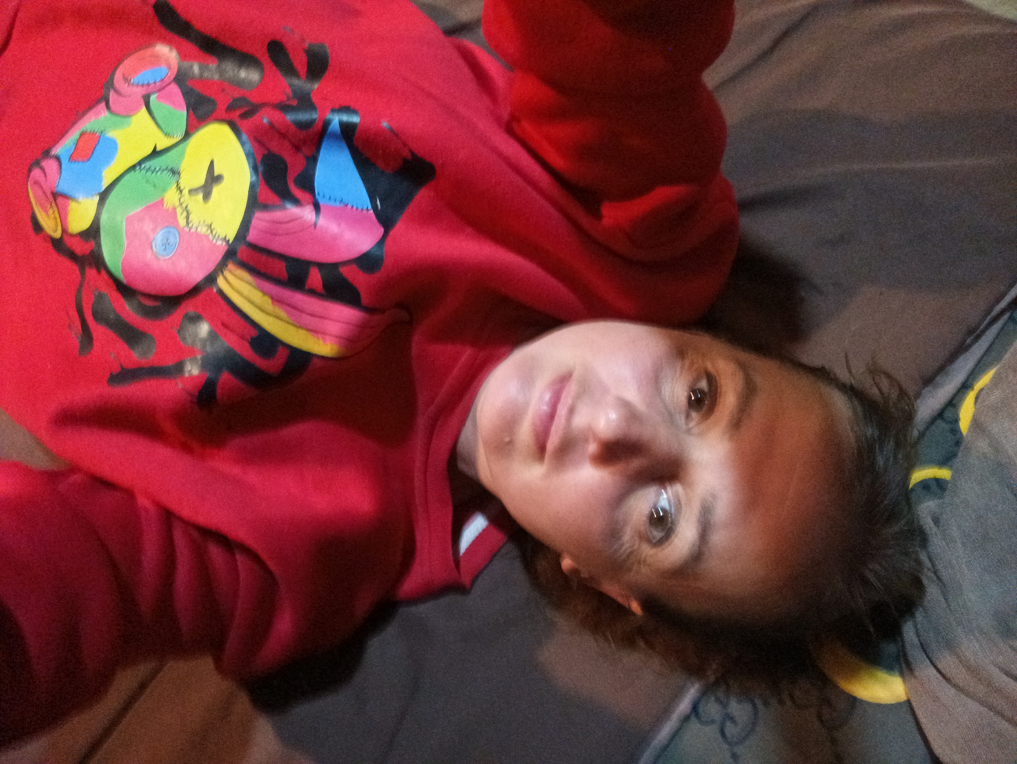 A woman is lying on a grey and blue patterned bedspread, wearing a red sweatshirt with a colorful cartoon design on the front. The location is Ban Tak Daet, Thailand.