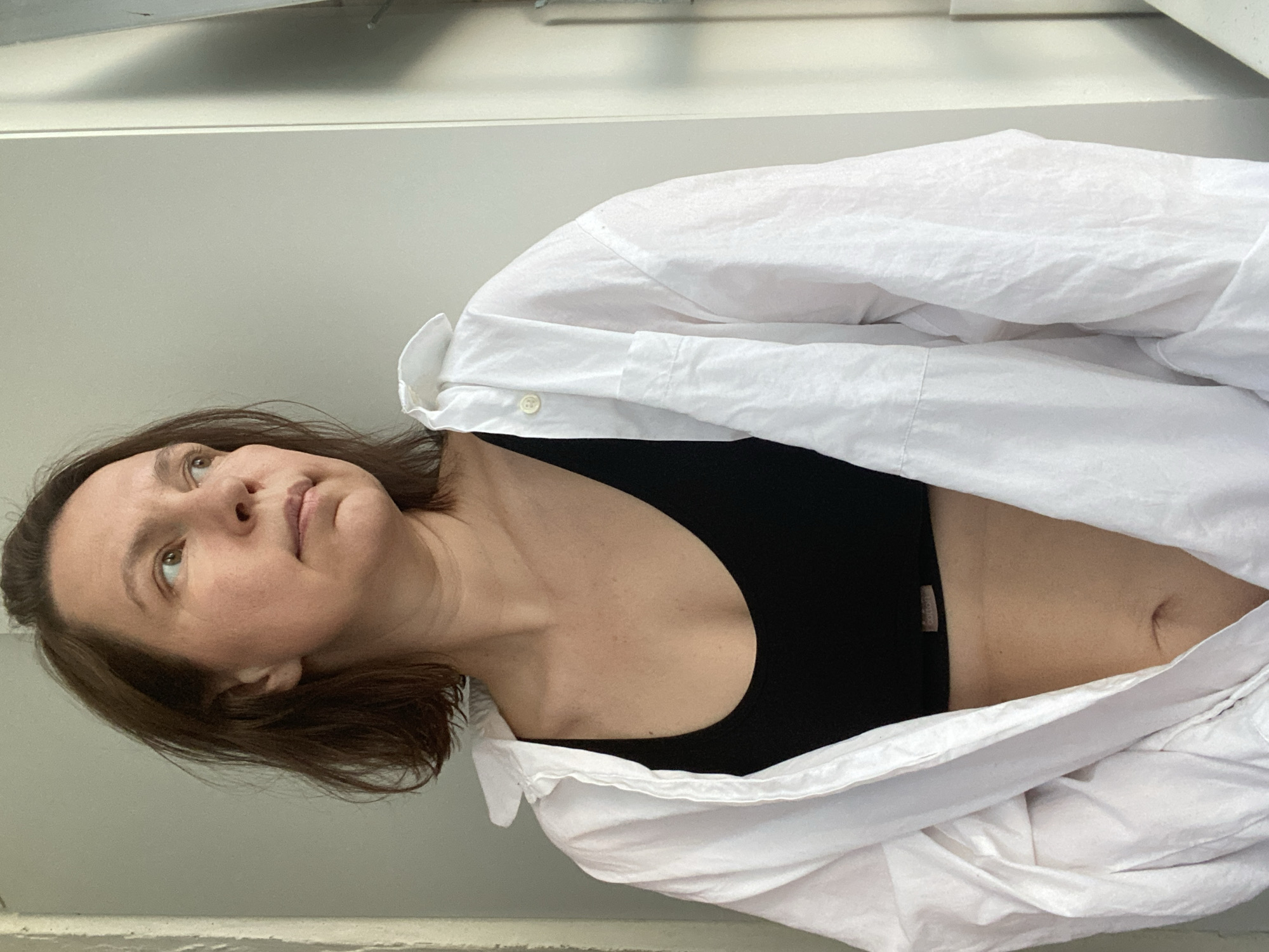 A woman with brown hair and fair skin is looking upwards. She is wearing a black sports bra and a partially open white collared shirt. The background appears to be a plain wall. There is no verifiable information to place the location of the image in Chaikovsky, Russia.