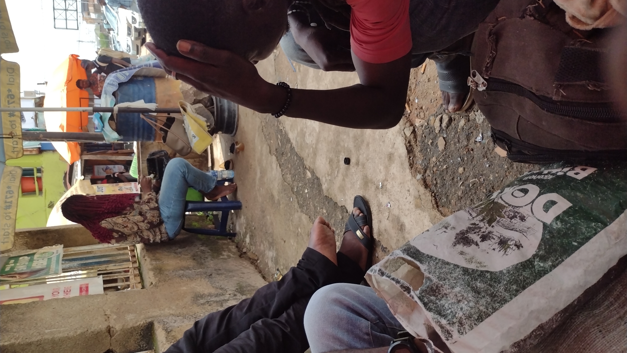 This media file captures a scene in Jos, Nigeria, focusing on several individuals and their surroundings. In the foreground, a person wearing black pants and blue jeans is visible, with their feet in black sandals. To their left, another individual is seated, wearing a patterned top and blue jeans, with their feet bare. Further back, a person with dark hair, wearing a red shirt, is seen from the shoulders up, with their hand on their head.
The setting appears to be an outdoor area, possibly a market or roadside stall, with various objects and structures visible. An orange awning can be seen in the background, along with some wooden stalls. A dark-colored backpack is also present in the scene, and a green and white bag with some text is in the foreground. The ground is paved with concrete and some gravel.
The overall impression is a moment of rest or pause in a busy environment. The lighting suggests it is daytime, likely bright, though the sky is not clearly visible. The atmosphere seems casual and informal, with people in everyday attire.
There is some text visible on signs and the green and white bag. One sign in the background has "Dial" printed on it in a vertical orientation. The green and white bag in the foreground has the word "Do" prominently displayed, along with other unreadable text and imagery. The scene likely depicts a typical day in a Nigerian urban setting.