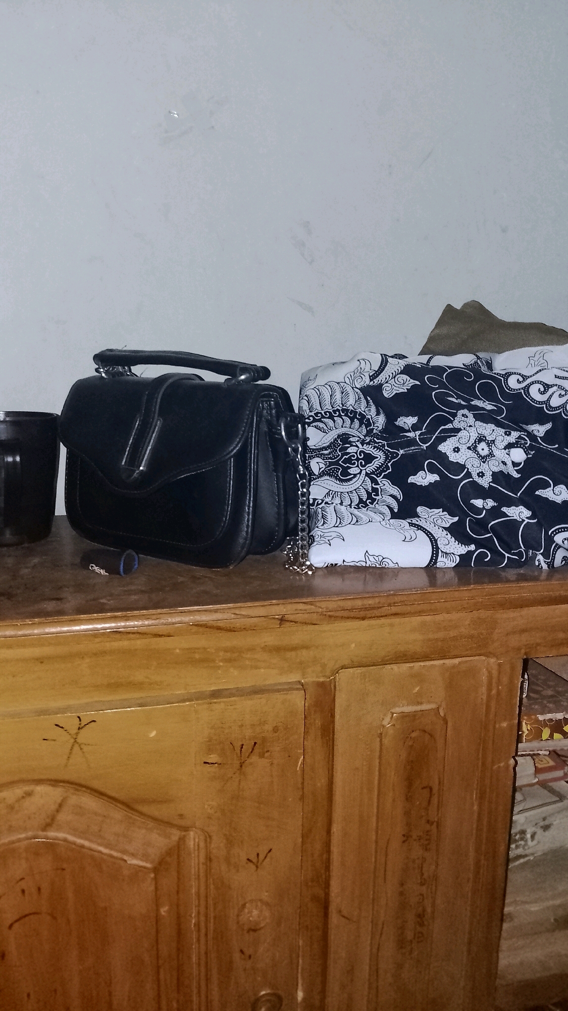A black leather purse with a chain strap sits on top of a wooden cabinet. To the right of the purse is a folded fabric with a black and white pattern. A black mug is visible to the left of the purse. The cabinet is made of light-colored wood and has decorative carvings on its doors. In the background, a plain white wall is visible. The scene appears to be indoors, likely in a domestic setting in Mulyo Haji, Indonesia.