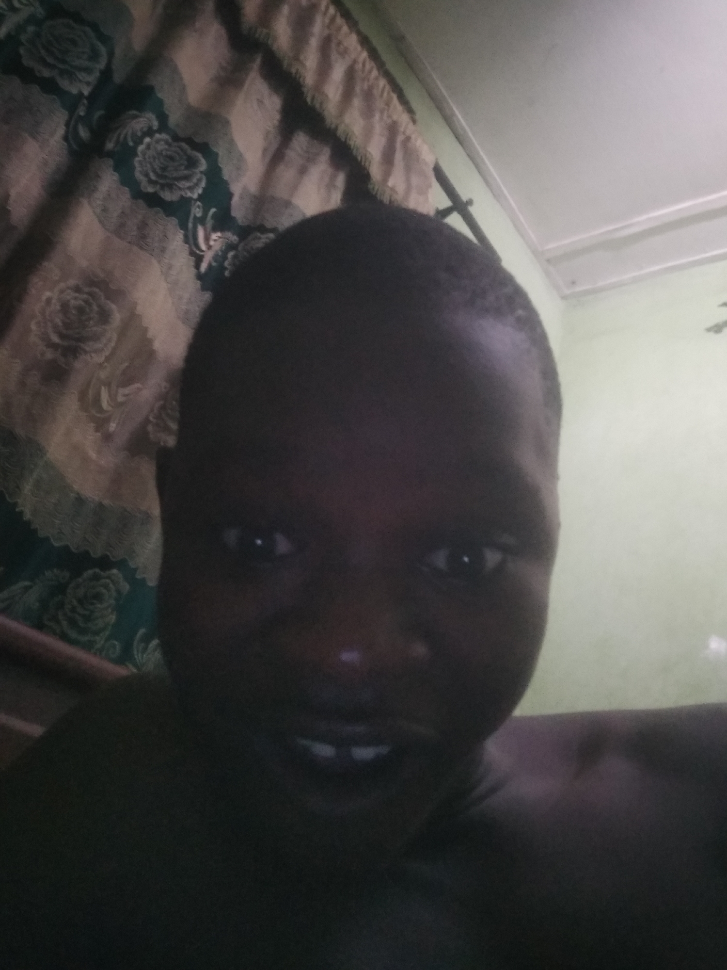 This dimly lit, close-up portrait features a dark-skinned individual, likely male, from Shangisha, Nigeria, looking directly at the camera. The person's face occupies a significant portion of the frame, captured in what appears to be a selfie or a very close-range photograph. Their eyes are wide open, and their mouth is slightly agape, revealing two front teeth. The individual appears bare-chested or is wearing a dark garment that blends into the shadows.
To the person's left (viewer's right), a patterned fabric, resembling a curtain or bedsheet, is visible. This fabric displays a dark green background adorned with a lighter floral (rose) pattern and bordered by wavy beige or light brown stripes. To the person's right (viewer's left) and above, a plain, light-colored wall or ceiling, possibly a light green hue, is partially seen, along with what could be molding or a light fixture. A dark, thin bar, potentially part of a bed frame or furniture, is visible in the lower-left corner of the image.
The low lighting suggests an indoor setting, possibly during nighttime or in a poorly illuminated room. The person's expression is ambiguous, hinting at curiosity or a slight, unenthusiastic smile. No text is visible within the image. The overall scene depicts a casual, intimate moment captured in a home environment.