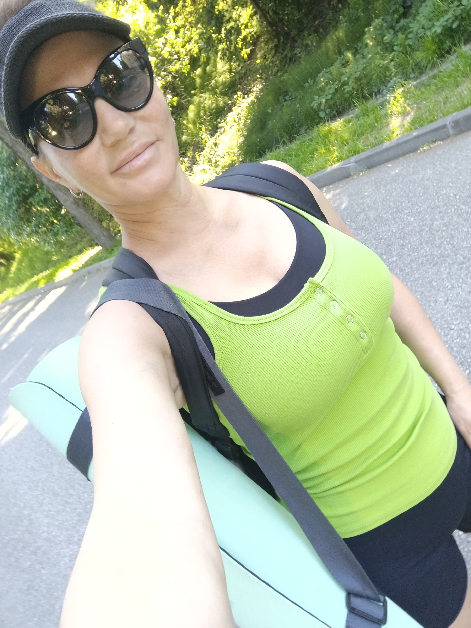 An outdoor, slightly elevated, waist-up, selfie of a woman wearing sunglasses, a dark hat, and a bright green ribbed tank top over a black sports bra. She is carrying a rolled-up light blue yoga mat over her left shoulder, with a dark grey strap from a backpack crossing her chest. The background is out of focus and shows a paved path next to a grassy area with trees. The sun is casting shadows.