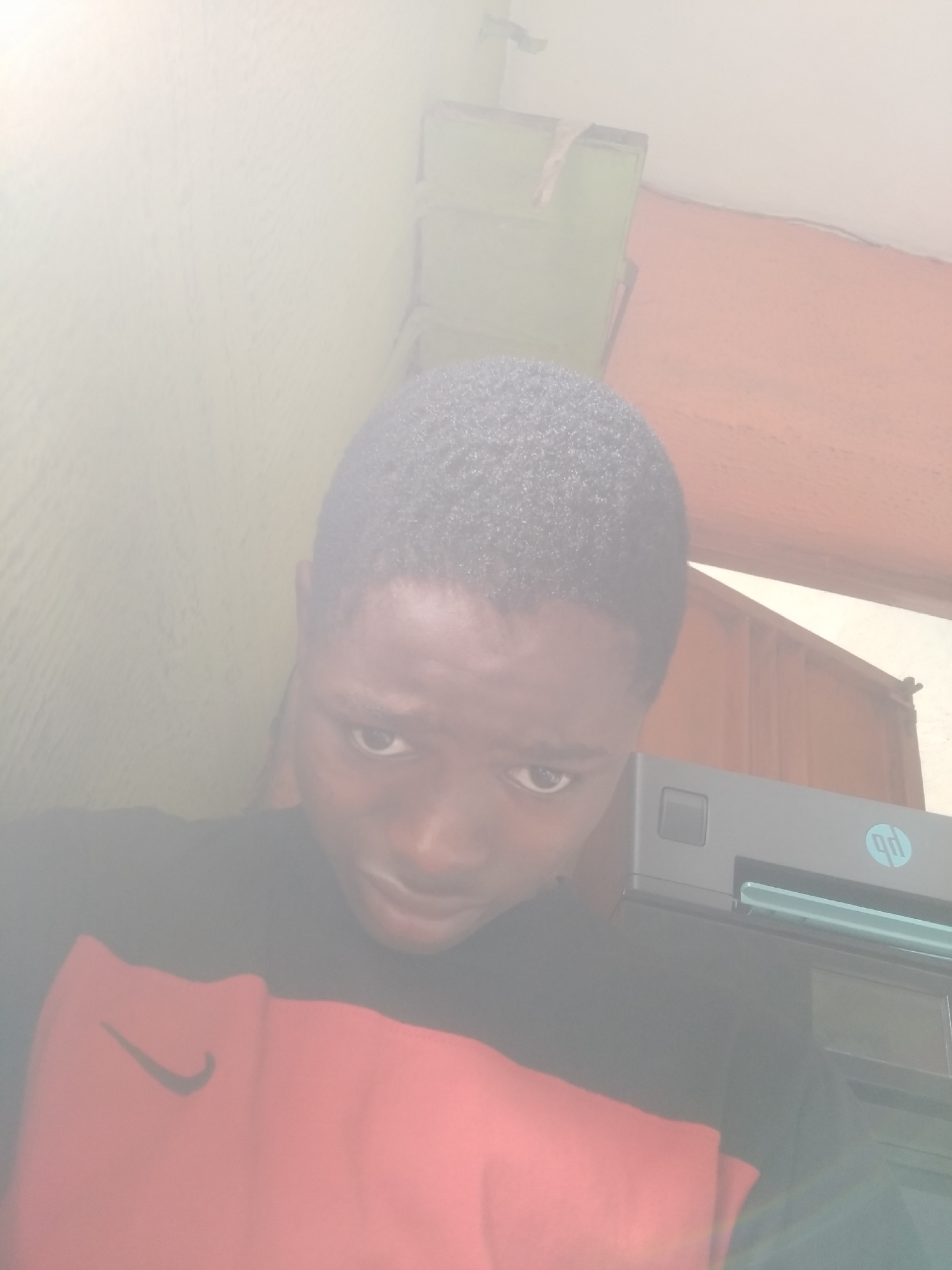 A young person, likely male, is featured in a brightly lit, somewhat overexposed portrait shot taken indoors in Shangisha, Nigeria. The individual has dark skin and short, dark, curly hair, looking directly at the camera with a neutral to slightly serious expression. They are wearing a black t-shirt with a prominent red section across the chest, which features a white Nike swoosh logo.
In the background, to the right of the person, a dark grey or black HP printer or scanner is visible, displaying its light blue 'hp' logo within a circle. The surrounding environment suggests an indoor room or office. Various wall sections are discernible: a very bright, almost white area dominates the upper left, a reddish-brown section is visible on the upper right, and a faint green element appears in the upper left background. The intense lighting, particularly on the left side, leads to some overexposure, making specific details and the exact time of day difficult to ascertain, though it suggests a well-lit environment, possibly daytime. No specific activity beyond posing for the picture is apparent.