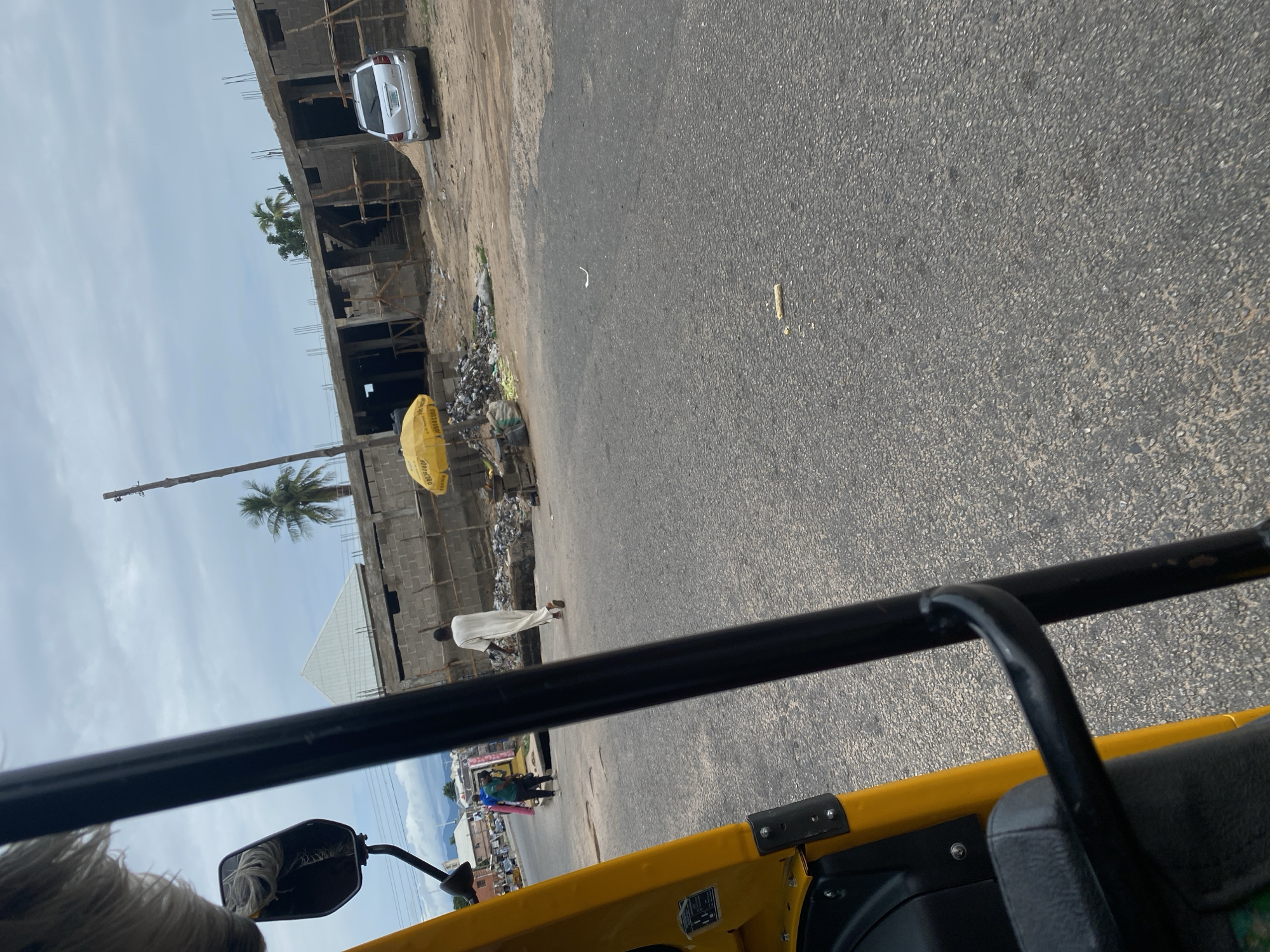 The image captures a street scene in Jalingo, Nigeria, from the perspective of someone in a yellow vehicle, likely a tuk-tuk. In the foreground, the yellow body of the vehicle and its black frame are visible, along with a side mirror reflecting some white cords.
The midground shows a paved road with some debris on the side, adjacent to a construction site. The building under construction appears to be multi-story, with exposed concrete blocks and wooden scaffolding. A white car is parked near the building. A person wearing a white robe is walking away from the camera on the side of the road, near a bright yellow umbrella. Further down the road, a few other individuals are visible, seemingly engaged in daily activities. Palm trees are interspersed within the scene, suggesting a tropical or subtropical climate.
The sky is overcast with some clouds, indicating a potentially humid or rainy day, though the light suggests it is daytime. The overall impression is of a busy, developing urban environment with elements of everyday life continuing amidst construction. No specific text is legible in the image.
