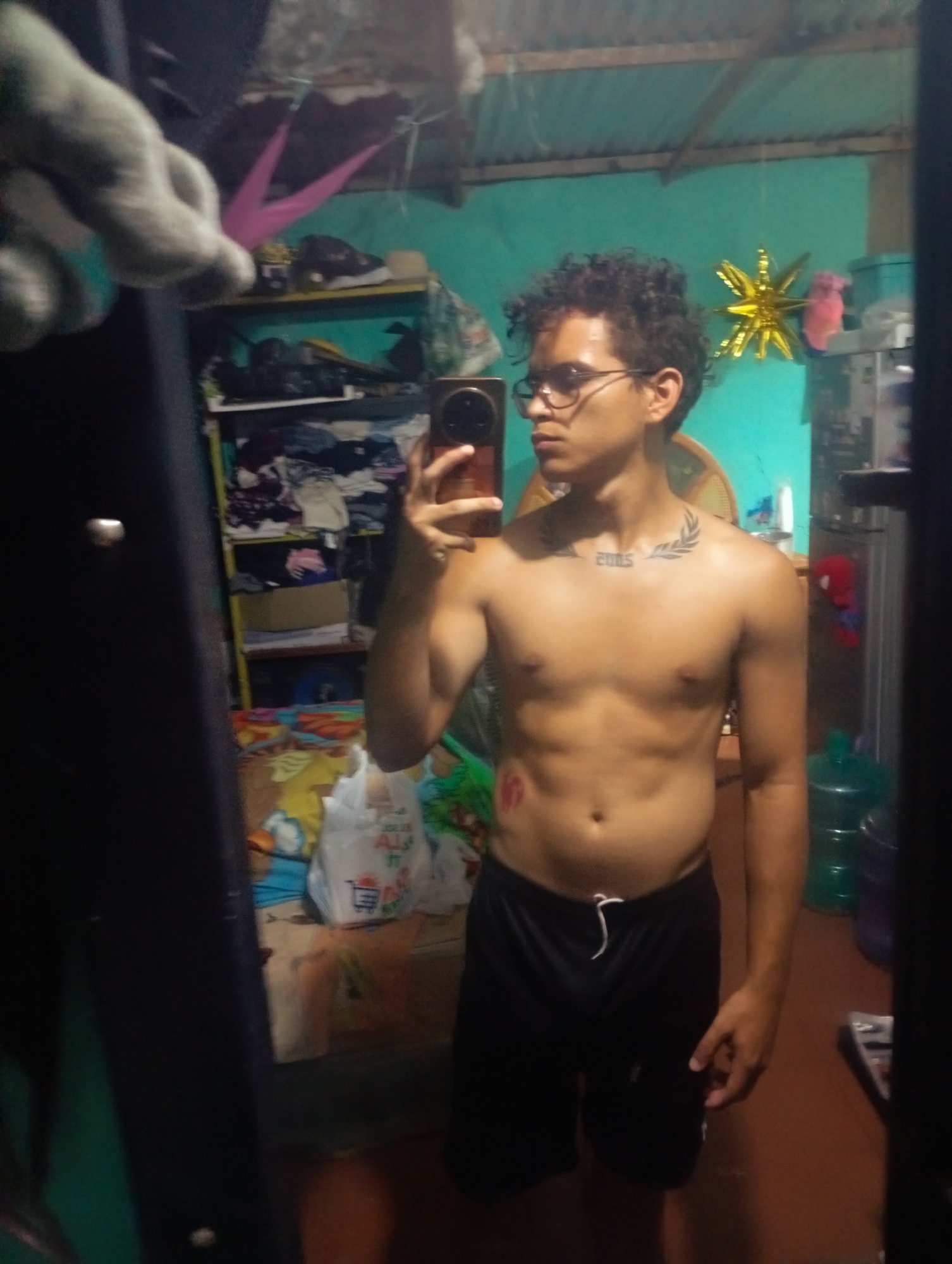 A young man with curly dark hair and glasses is taking a mirror selfie in a room. He is shirtless, wearing black shorts with a white drawstring. A tattoo of a laurel wreath and "2005" is visible on his chest. To his left, a red lipstick mark is on his skin. In the background, shelves are filled with clothing and various items. A large, clear bottle of water is visible on the right side of the image. The walls are painted a teal color. The ceiling appears to be made of corrugated metal. The overall impression is that of a casual, possibly domestic setting. The presence of the water bottle and the general state of the room suggest it could be a home or a similar informal dwelling in Altagracia, Venezuela.